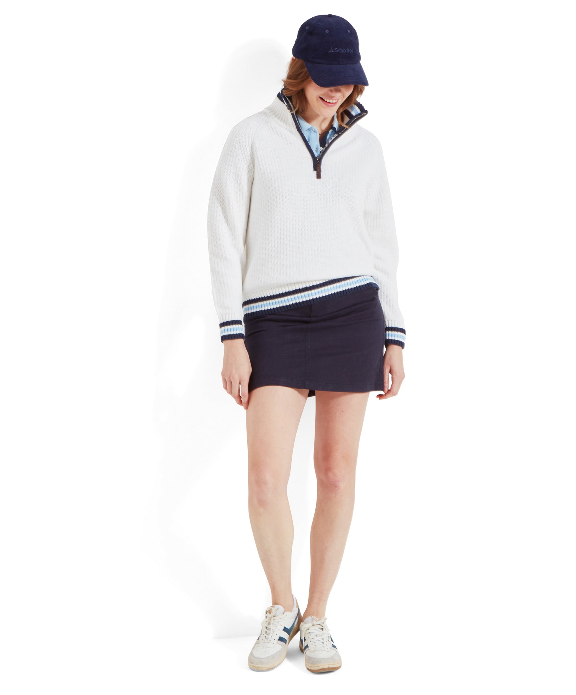 The same woman is standing, showcasing the full outfit which includes a Schöffel Havelet Bay 1/4 Zip Jumper for Women in Blue, a navy blue mini skirt, white trainers, and a navy blue cap. She is looking down and smiling.