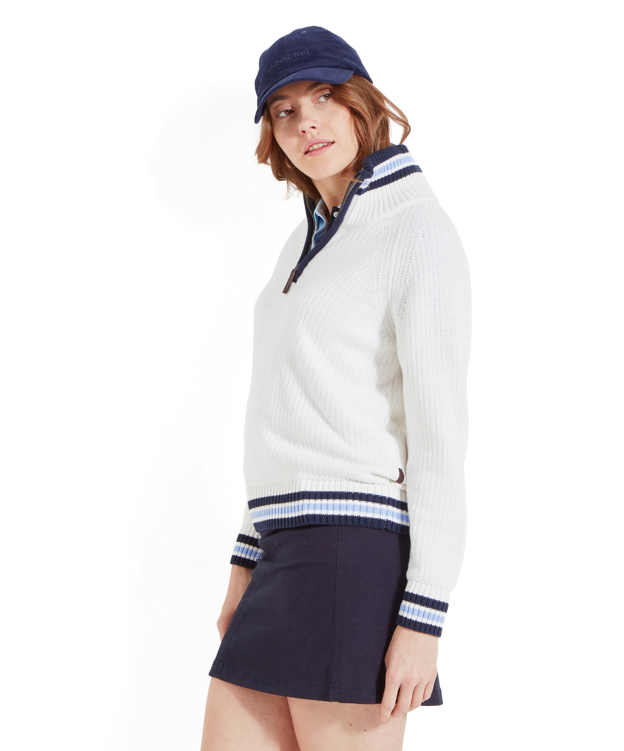 The woman is shown from a side angle, highlighting the quarter-zip feature of the white jumper with blue and white striped accents. She is also wearing a navy blue mini skirt and a navy blue cap.