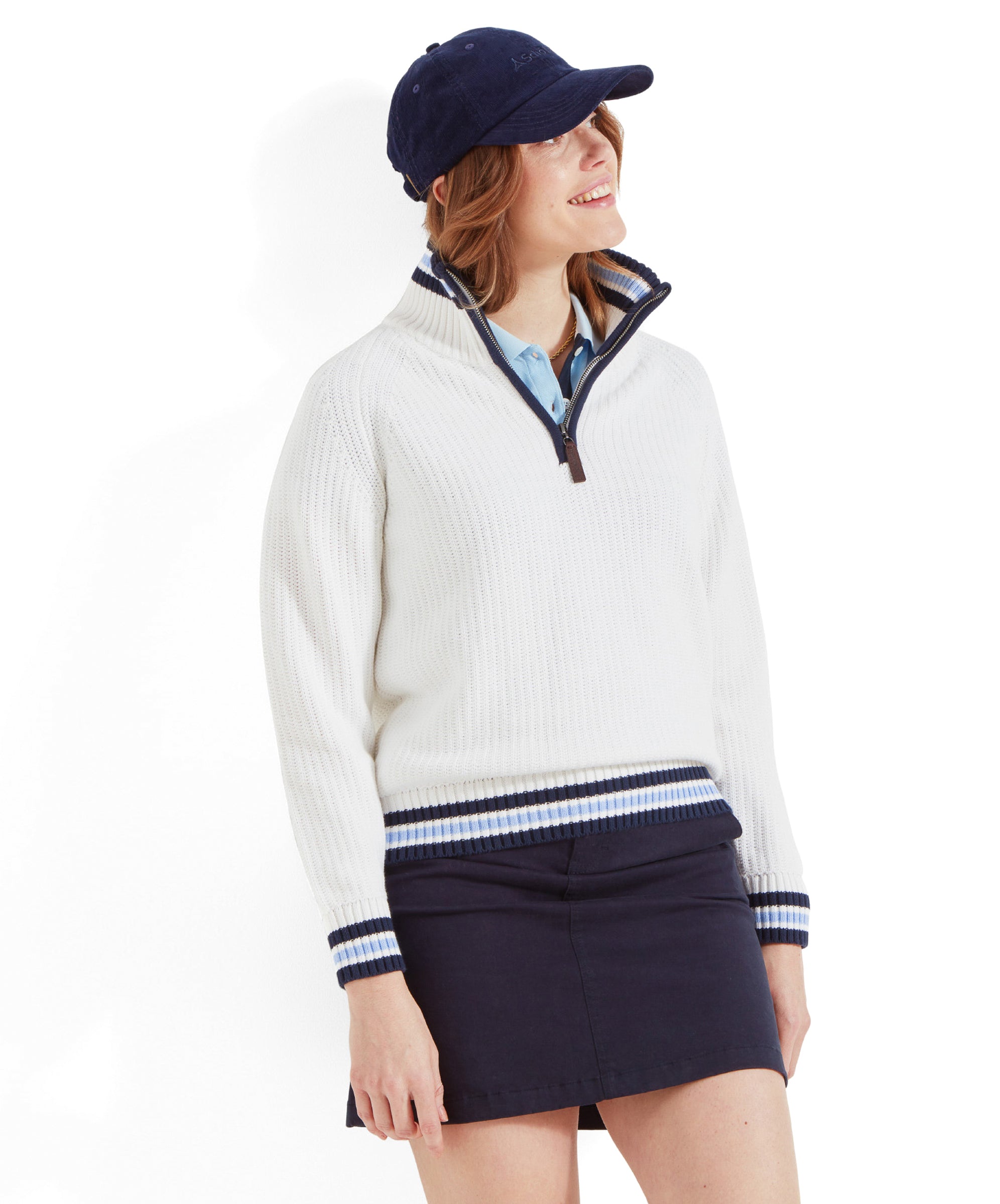 The woman is facing forward and smiling. She is wearing a Schöffel Havelet Bay 1/4 Zip Jumper for Women in Blue, a navy blue mini skirt, white trainers, and a navy blue cap.