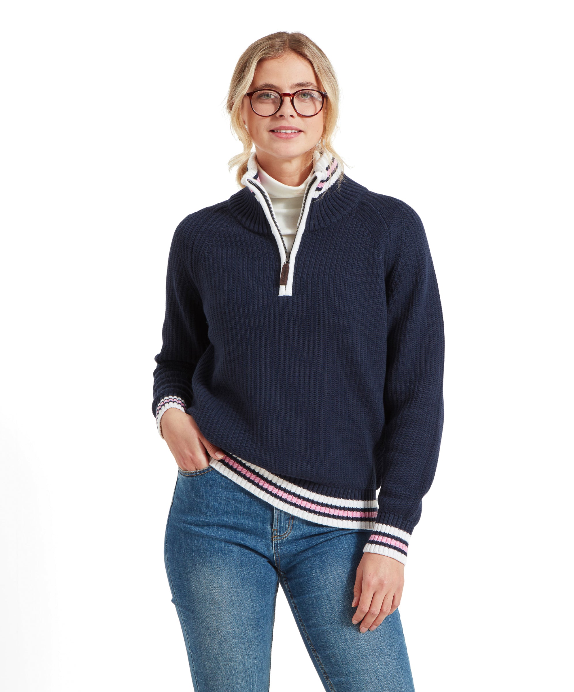 A woman with blonde hair and glasses wearing a Schöffel Havelet Bay 1/4 Zip Jumper for Women in Navy with white, pink, and navy striped accents on the collar, cuffs, and hem. She is also wearing a white turtleneck underneath and blue jeans.