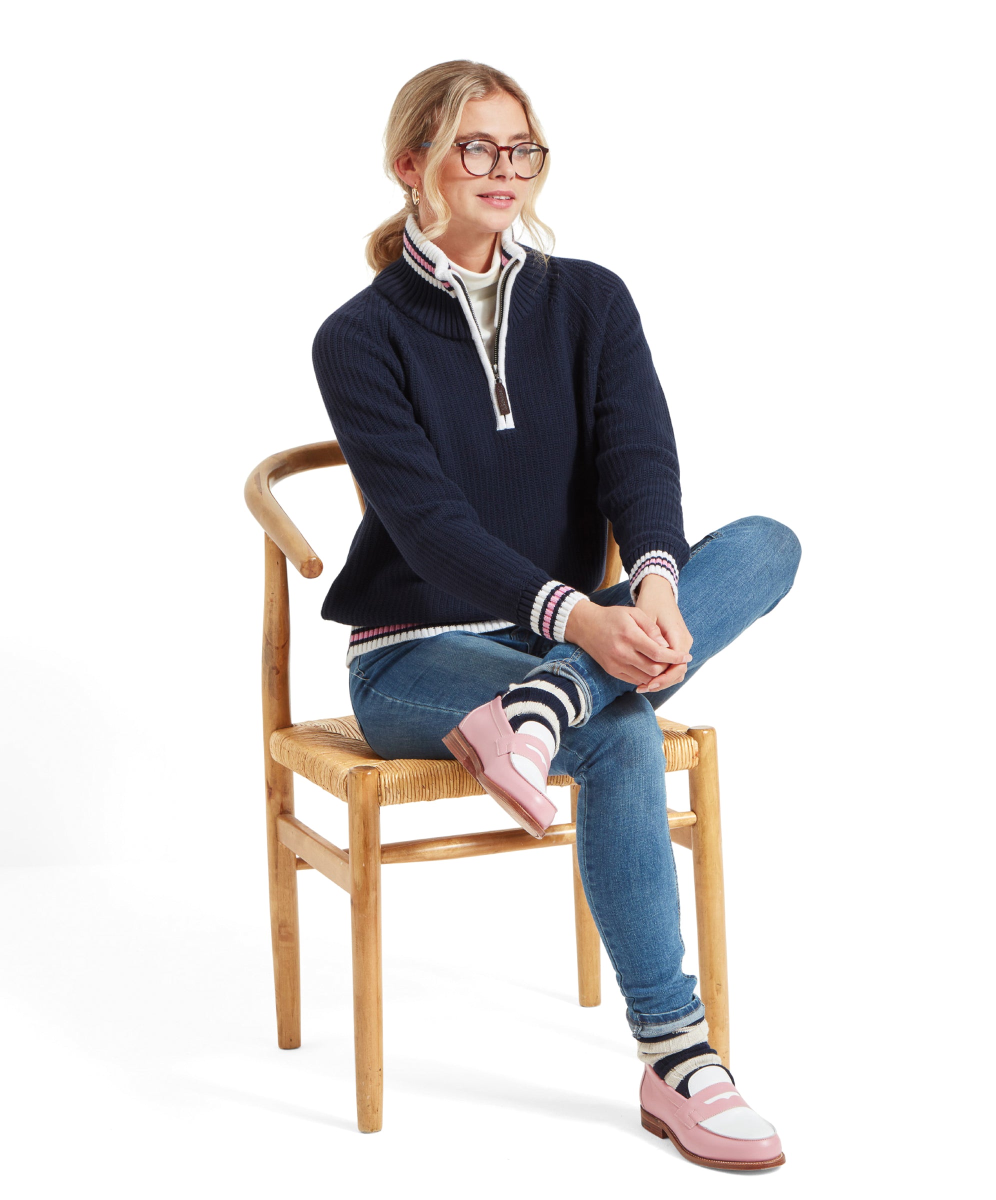 The same woman is sitting on a wooden chair with one leg crossed over the other. She is wearing a Schöffel Havelet Bay 1/4 Zip Jumper for Women in Navy, blue jeans, striped socks, and pink loafers with white details. She is smiling and looking to the side.