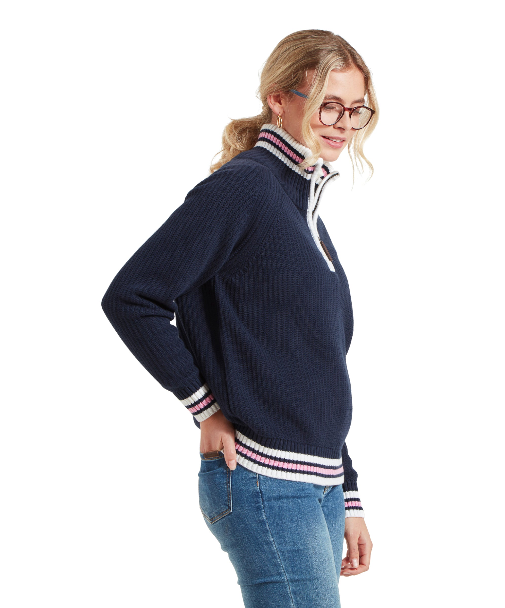 A side profile of the woman, highlighting the details of the Schöffel Havelet Bay 1/4 Zip Jumper for Women in Navy with white, pink, and navy striped accents. She is wearing blue jeans and her hair is tied back in a ponytail.