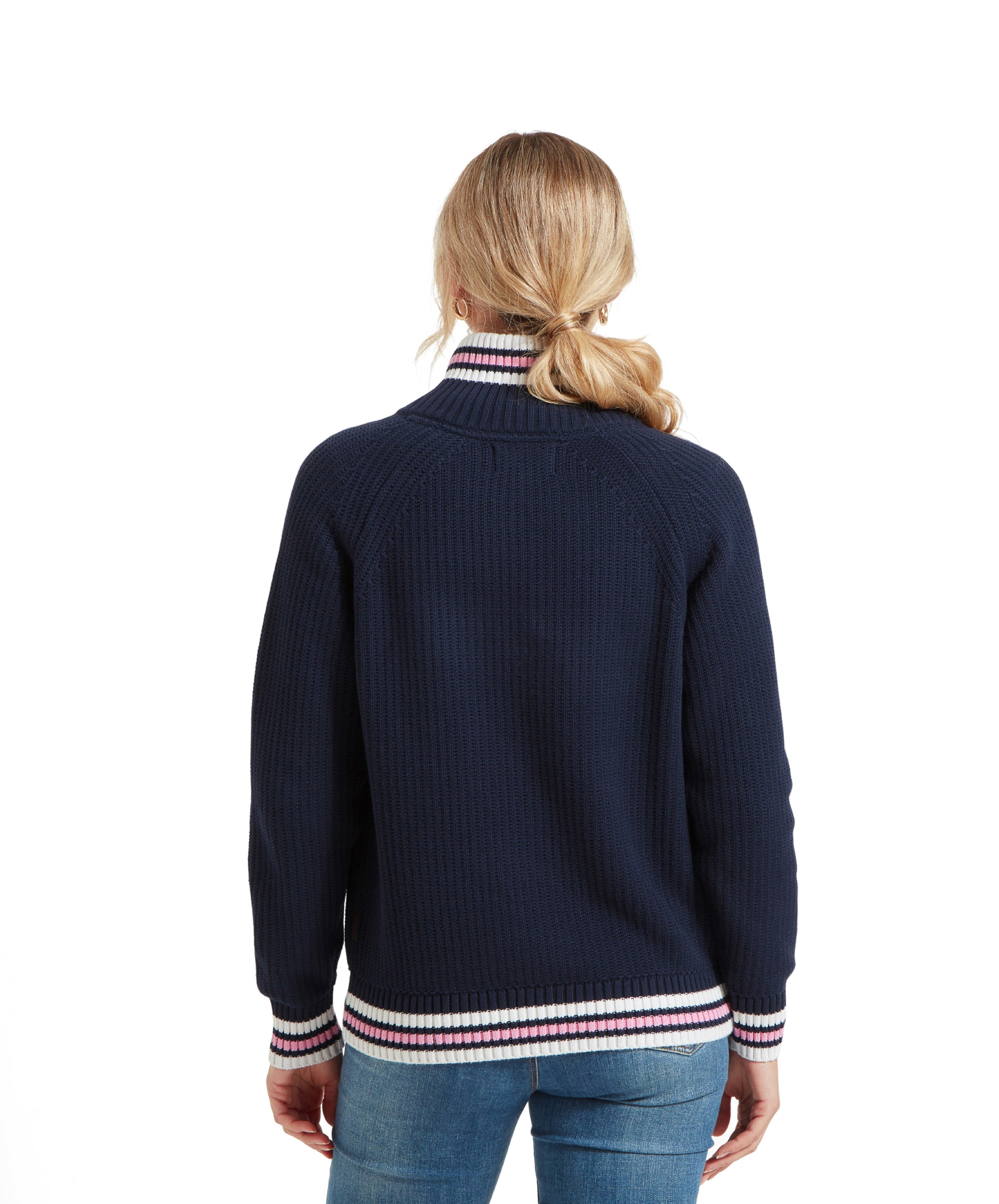 The woman is turned around, displaying the back of the Schöffel Havelet Bay 1/4 Zip Jumper for Women in Navy with white, pink, and navy striped accents on the collar, cuffs, and hem. She is wearing blue jeans and has her hair in a ponytail.