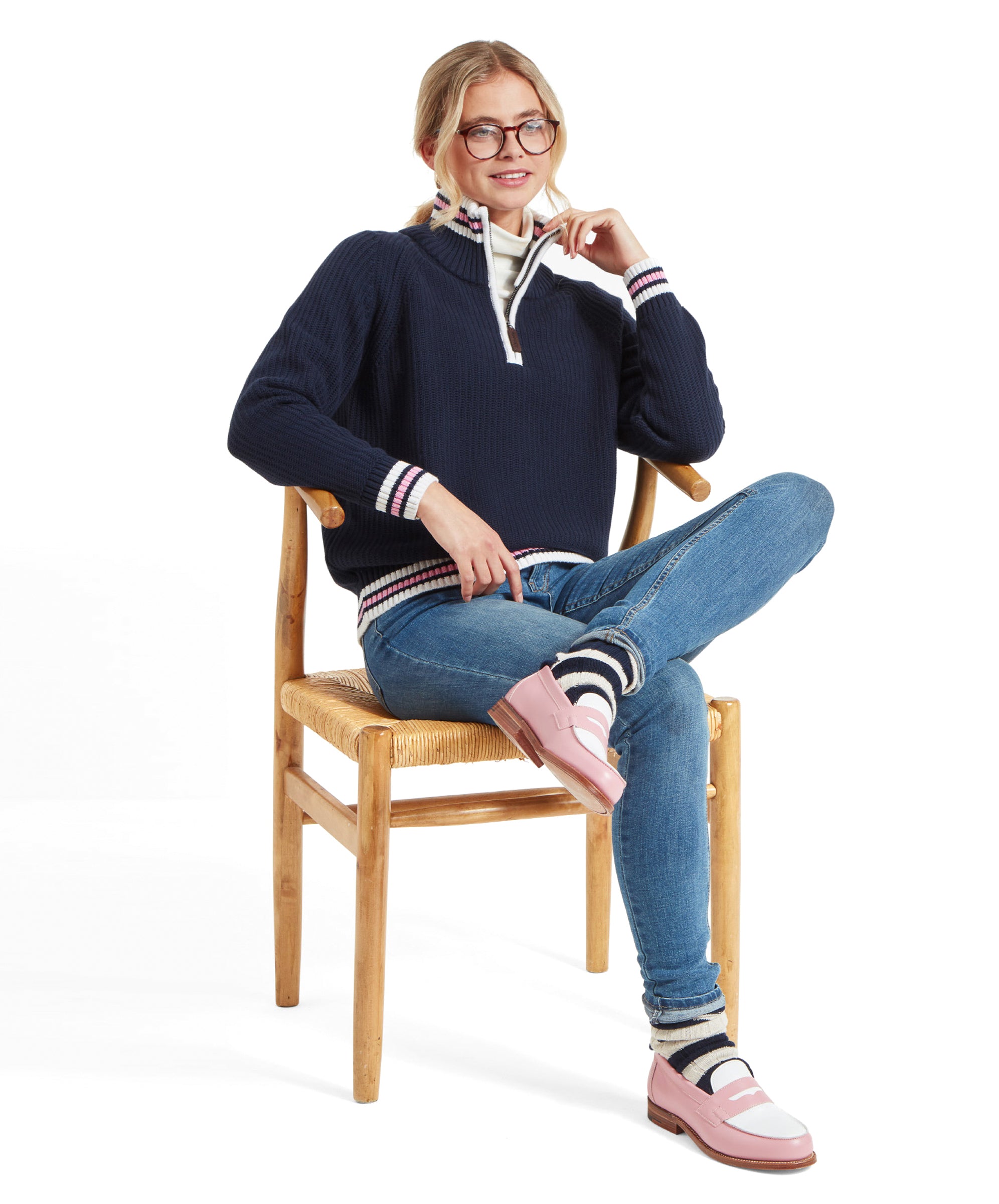 The woman is sitting on a wooden chair with one leg crossed over the other. She is wearing a Schöffel Havelet Bay 1/4 Zip Jumper for Women in Navy, blue jeans, striped socks, and pink loafers with white details. She is smiling and resting her elbow on the back of the chair.