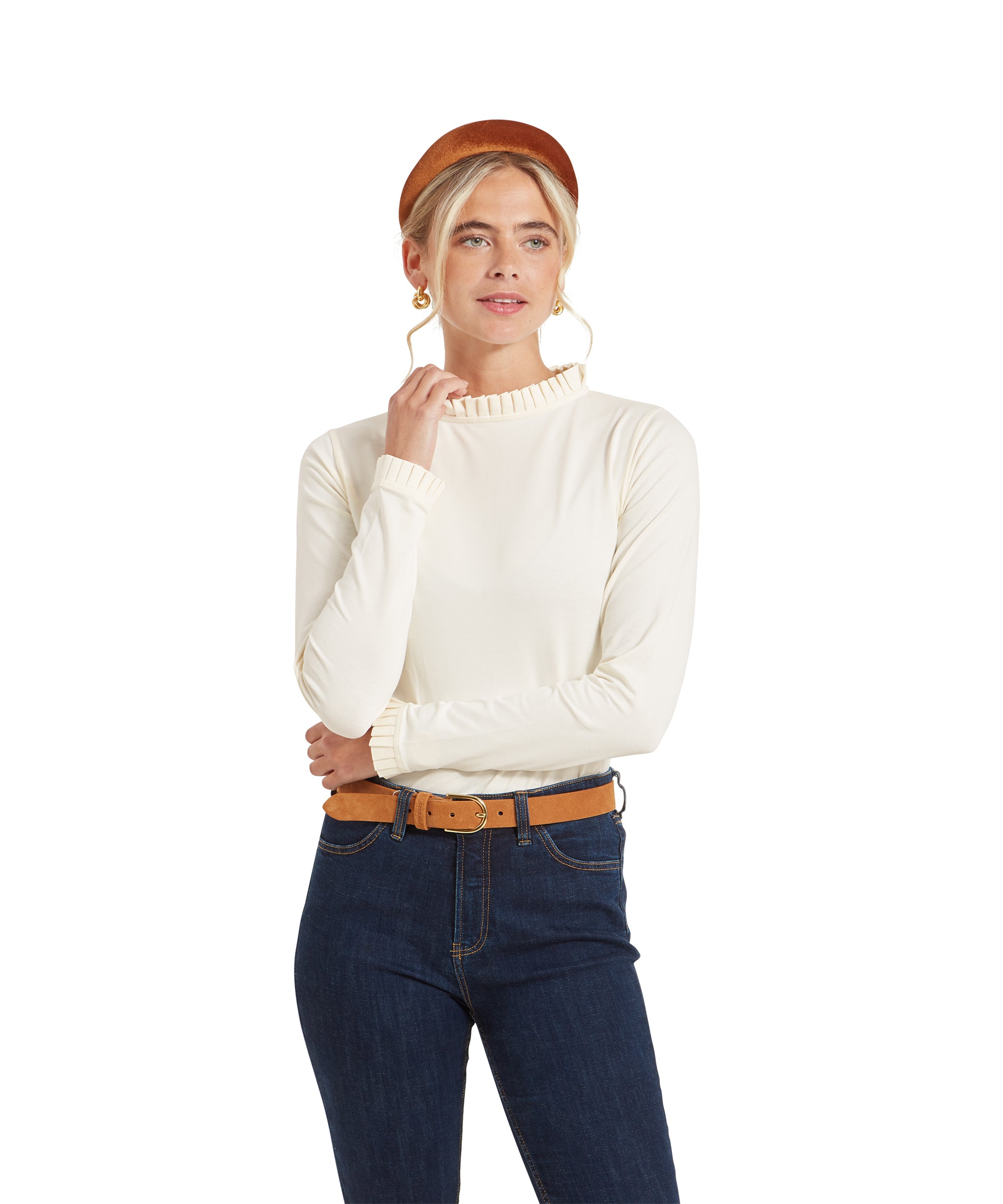 A woman wearing a Schöffel Ilkley Top for Women in Cream with a pleated high neckline and cuffs. She has blonde hair styled in an updo, wears gold hoop earrings, and is accessorised with a tan headband and belt. She is standing with one hand near her chin and smiling.