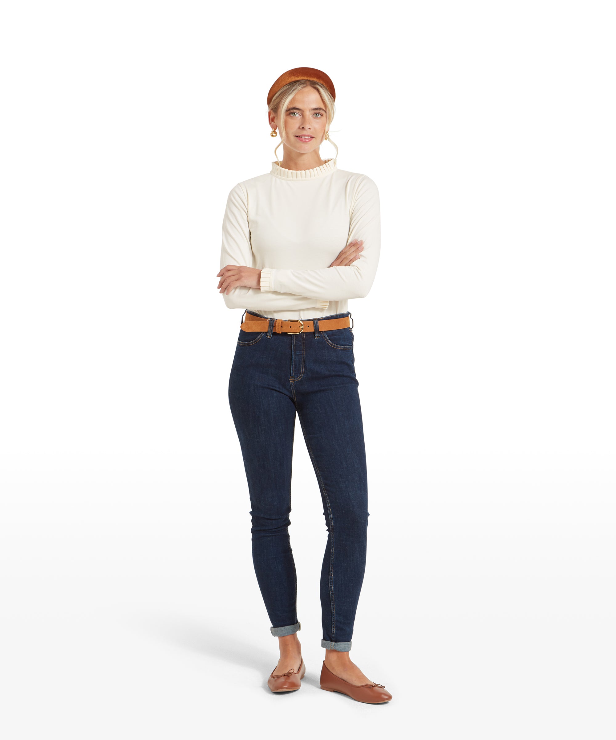 The same woman as in the previous image, shown in a full-body view. She is wearing the Schöffel Ilkley Top for Women in Cream with dark blue jeans, a tan belt, tan flats, and a tan headband. She is standing with her arms crossed.