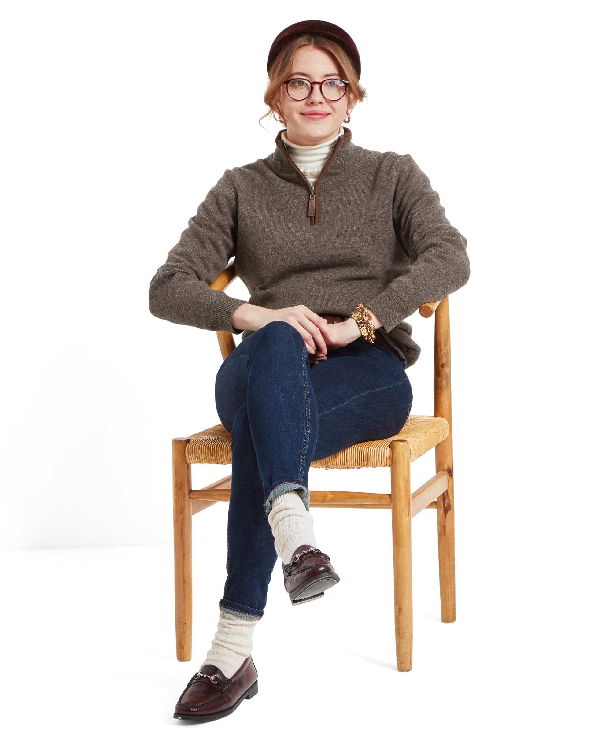 A woman sitting on a wooden chair wearing a Schöffel Women's Lambswool Aerobloc 1/4 Zip for Womens in Brown, dark blue jeans, and loafers with socks. She has a brown headband and glasses.