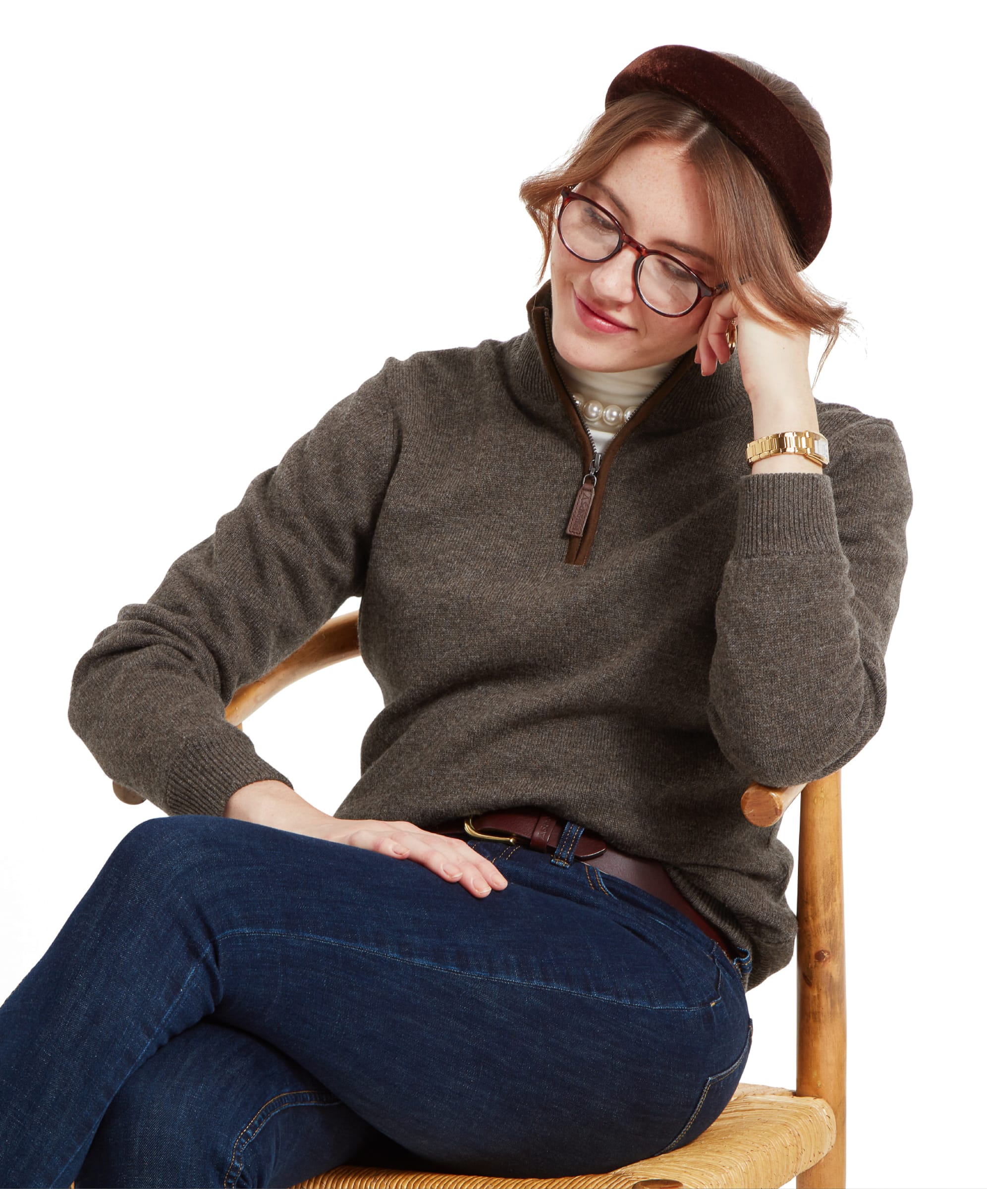 A woman wearing a Schöffel Women's Lambswool Aerobloc 1/4 Zip for Womens in Brown, dark blue jeans, and a brown headband. She is sitting with her head resting on her hand, smiling slightly.