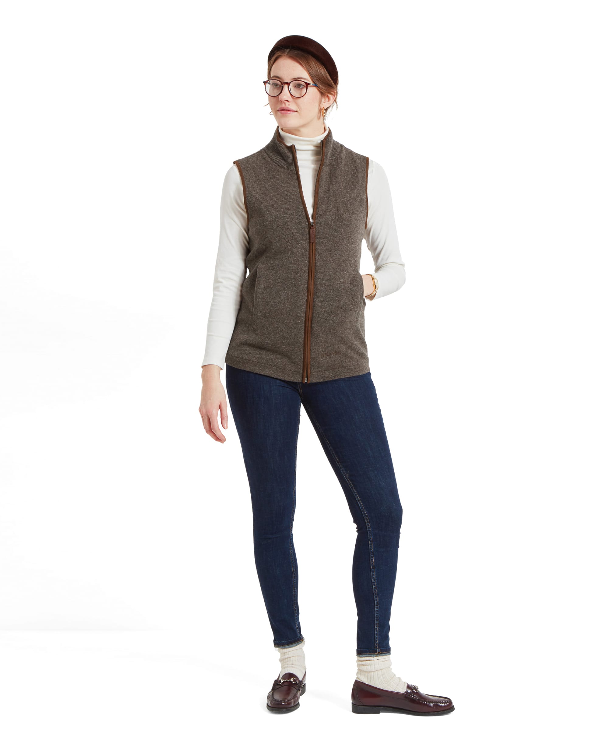 Full body image of a woman wearing a Schöffel Women's Lambswool Aerobloc Gilet for Womens in Brown over a white turtleneck, paired with dark jeans. She is standing casually with her hands in the pockets of the gilet