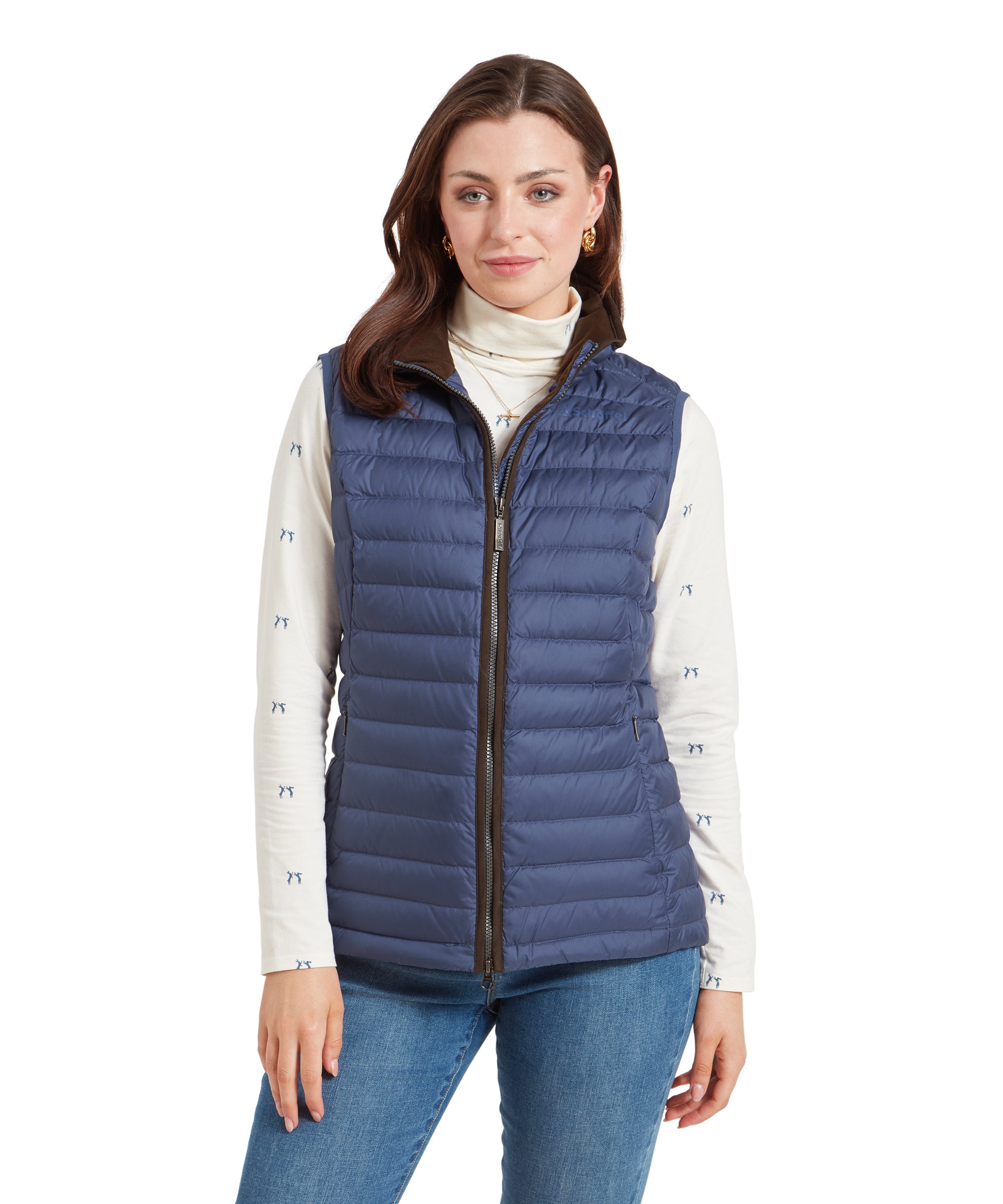 A woman wearing a Schöffel Launde Down Gilet for Women in Blue over a cream long sleeve top with a subtle print, paired with light denim jeans. She is smiling and standing with her hands held at her sides.