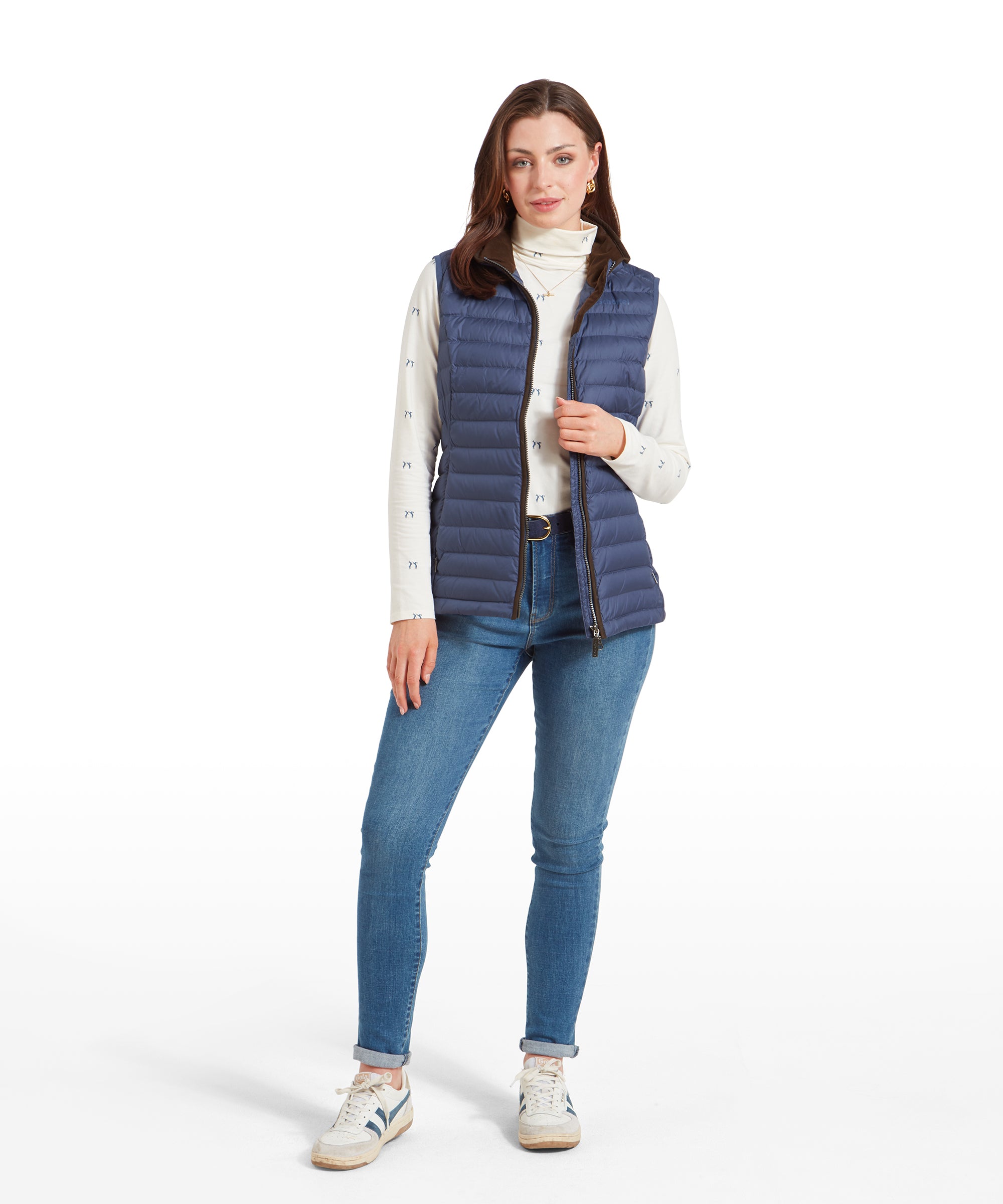 A full-body shot of a woman wearing a Schöffel Launde Down Gilet for Women in Blue over a cream long sleeve top with a delicate pattern, paired with denim jeans and white and blue trainers. The gilet is worn open and the model is adjusting the front.