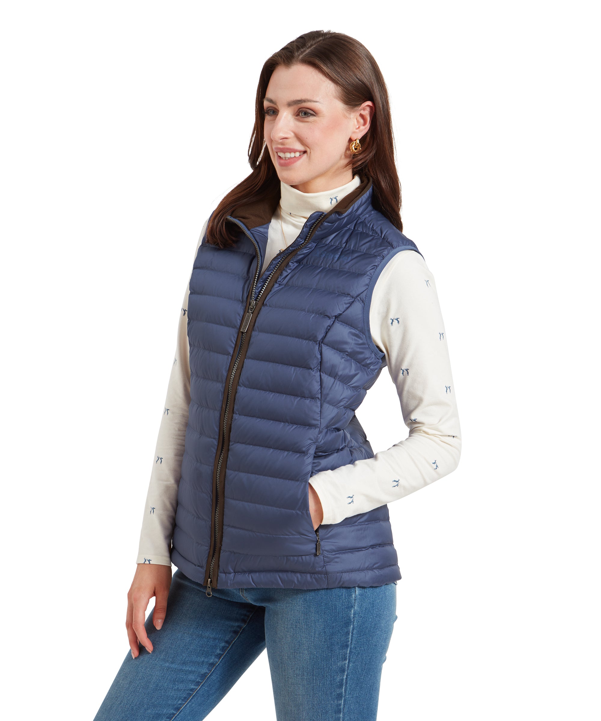 Side view of a woman wearing the Schöffel Launde Down Gilet for Women in Blue over a cream long sleeve top, paired with light denim jeans. The woman stands with her hand resting in a pocket. Her outfit is accessorised with gold hoop earrings.