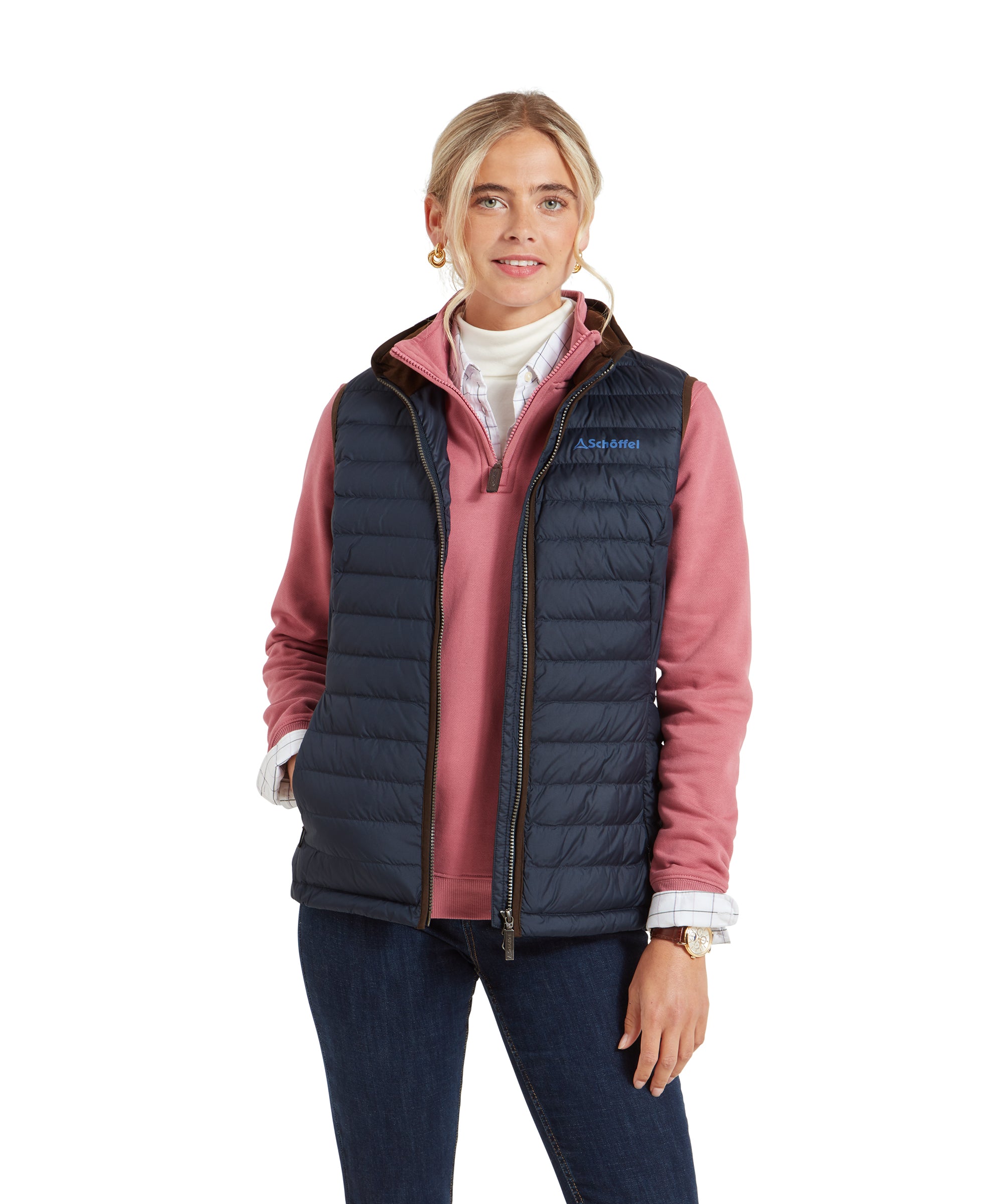 A woman is wearing a Schöffel Launde Down Gilet for Women in Navy with a pink mid-layer underneath and jeans. The gilet features a brown trim and the Schöffel logo on the left chest.