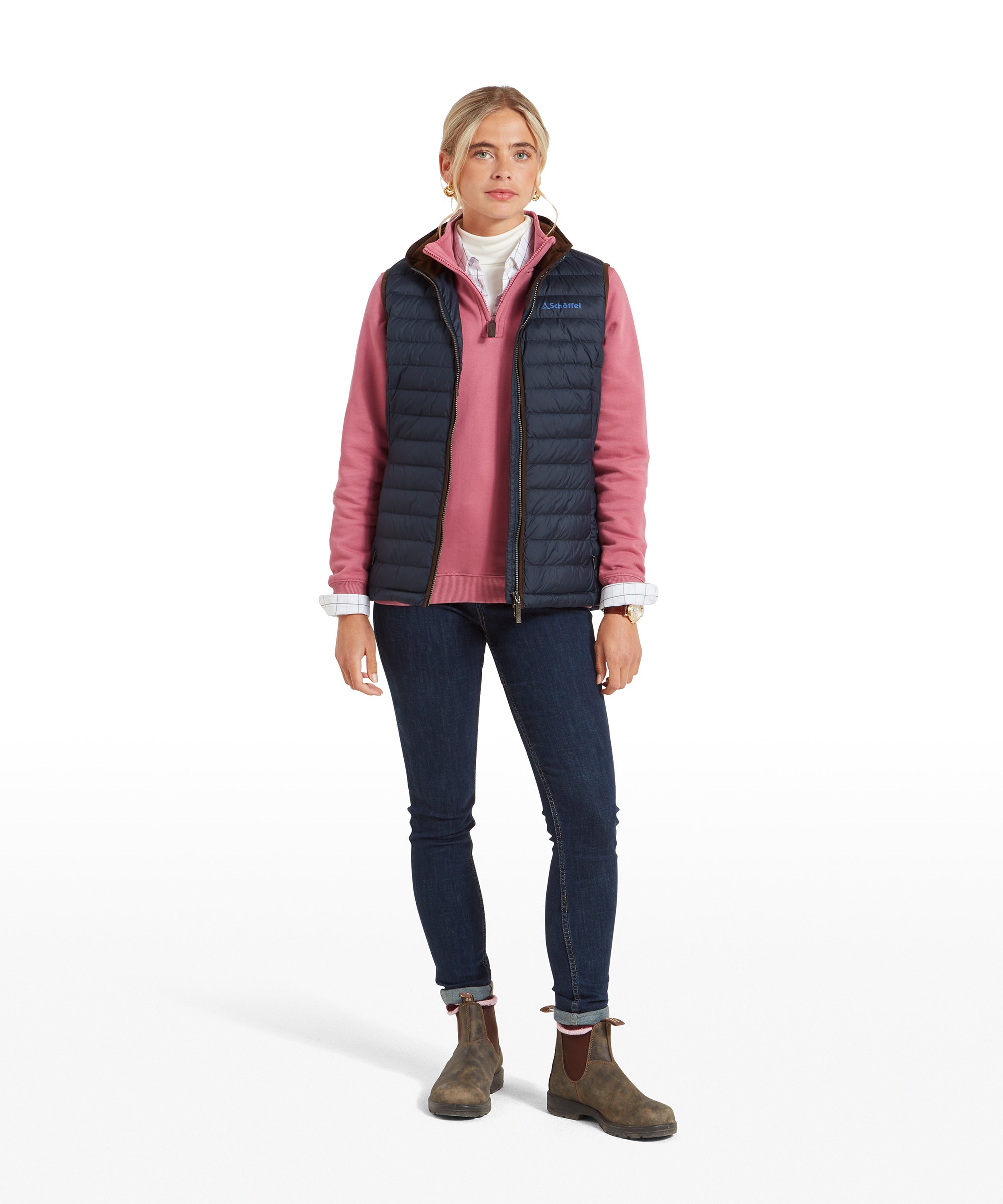 The woman is standing fully, showing the entire outfit with the Schöffel Launde Down Gilet for Women in Navy, pink mid-layer, jeans, and brown boots. The outfit has a casual and comfortable look.