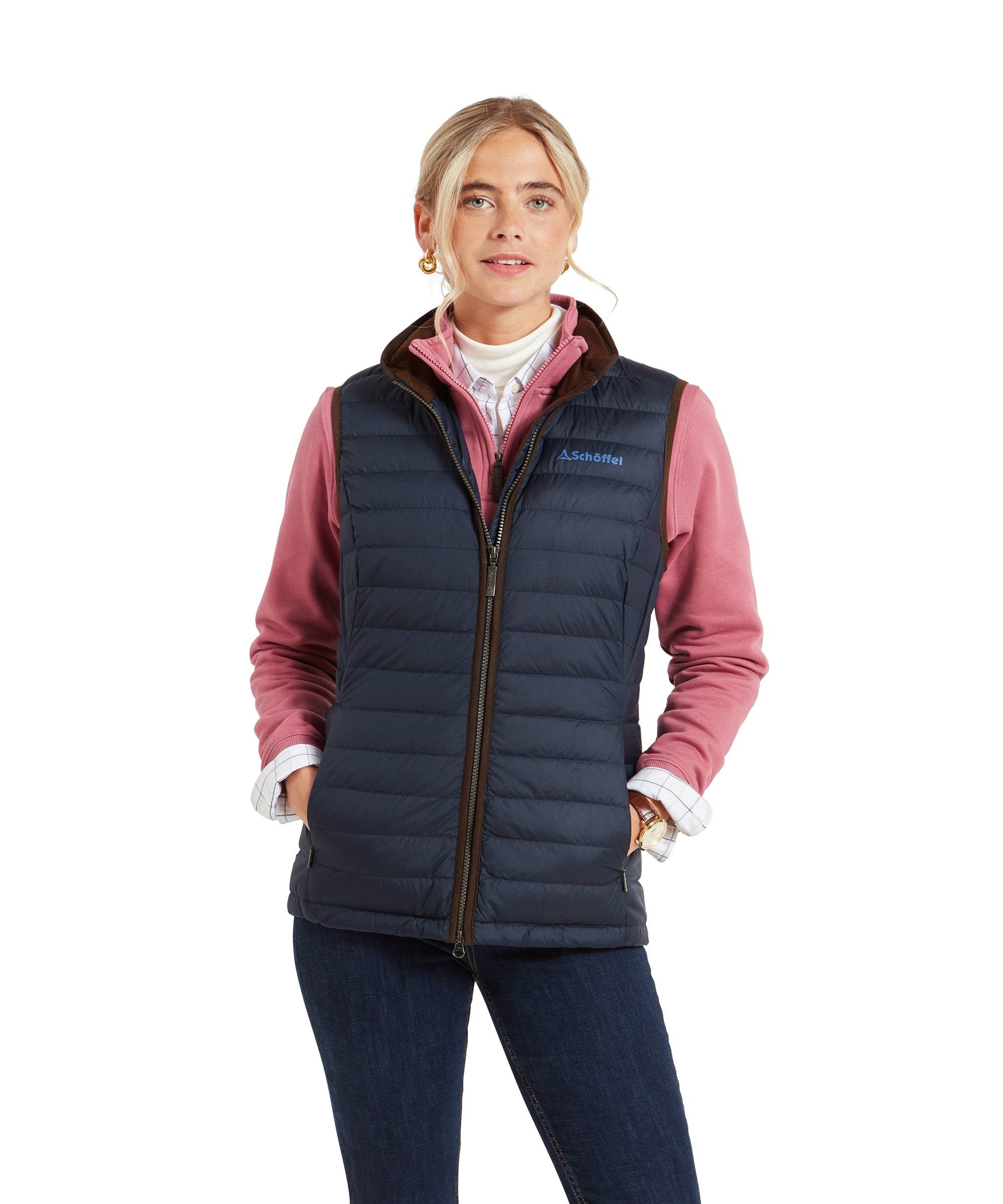 A woman wearing a Schöffel Launde Down Gilet for Women in Navy with brown trim over a pink mid-layer and white turtleneck. The gilet features the Schöffel logo in blue on the left chest. She is also wearing jeans and gold hoop earrings, with her blonde hair styled in a low bun.