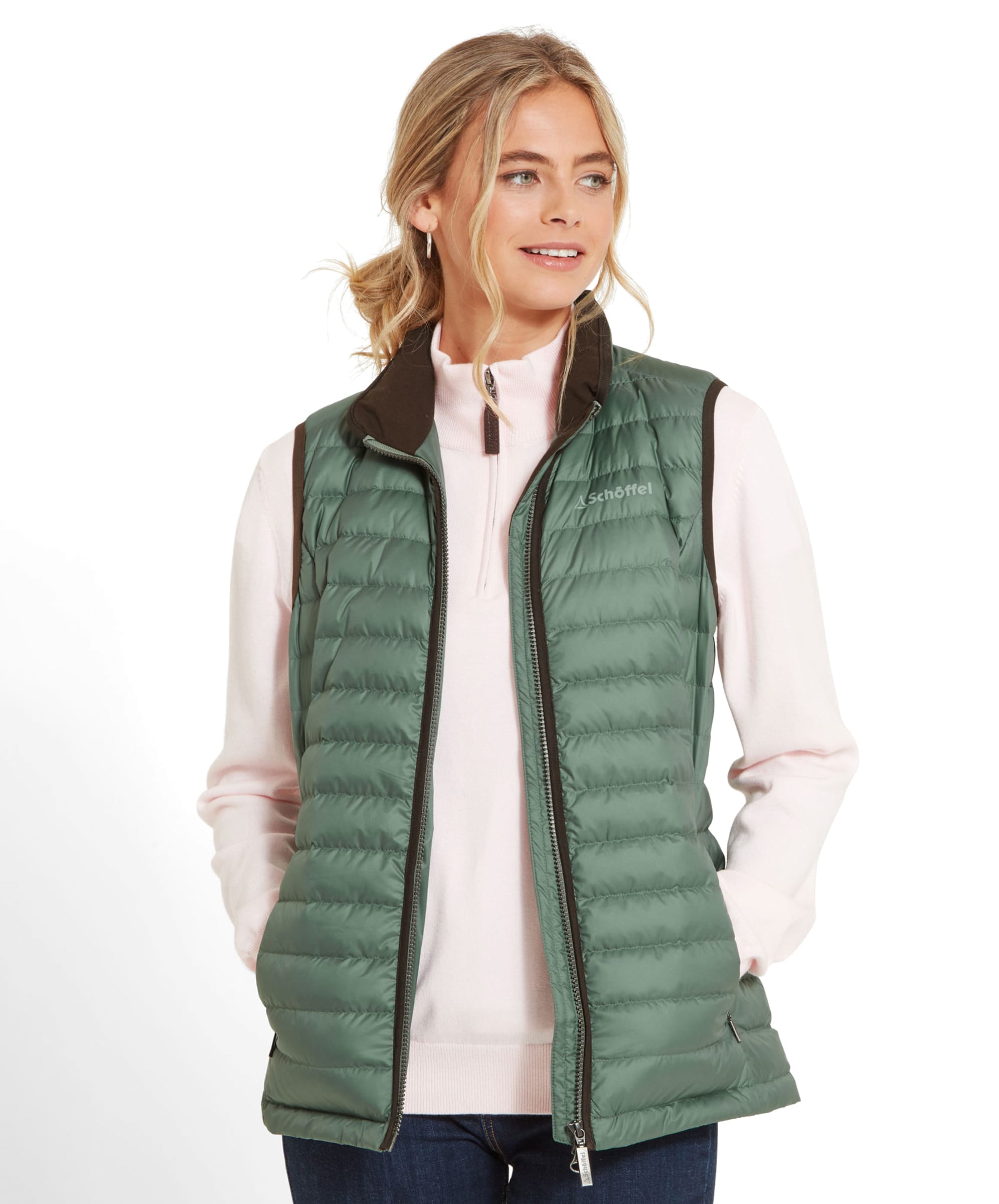 A woman wearing the Schöffel Launde Down Gilet for Women in Green over a pale pink 1/4 zip sweater and dark denim jeans. The woman is gently smiling and looking away and he hands are placed in the gilet pockets.