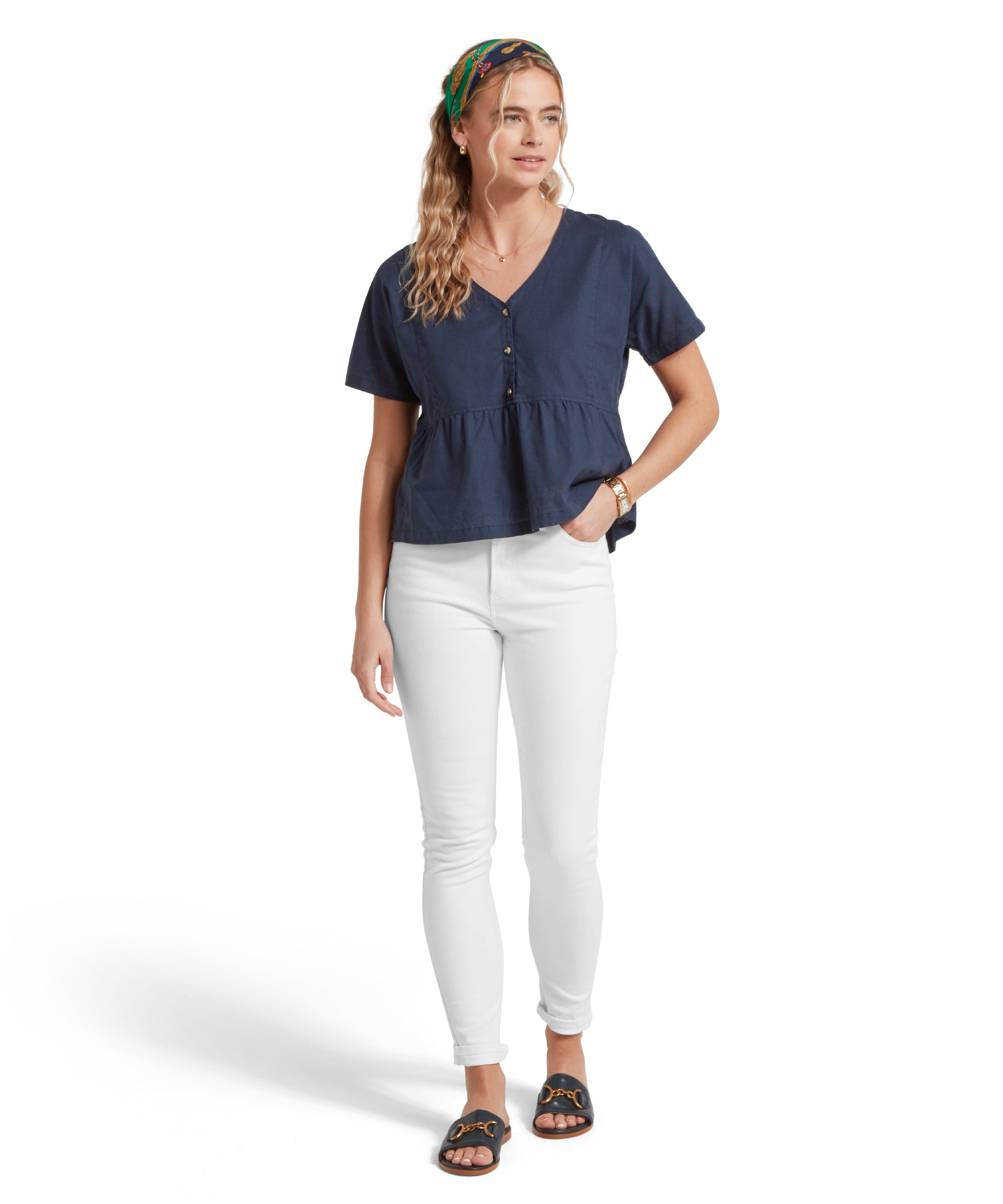 Full-height image of a female model wearing a v-neck navy linen Schöffel top, paired with white jeans and black sandals. She is standing in a relaxed pose with one hand in her pocket.