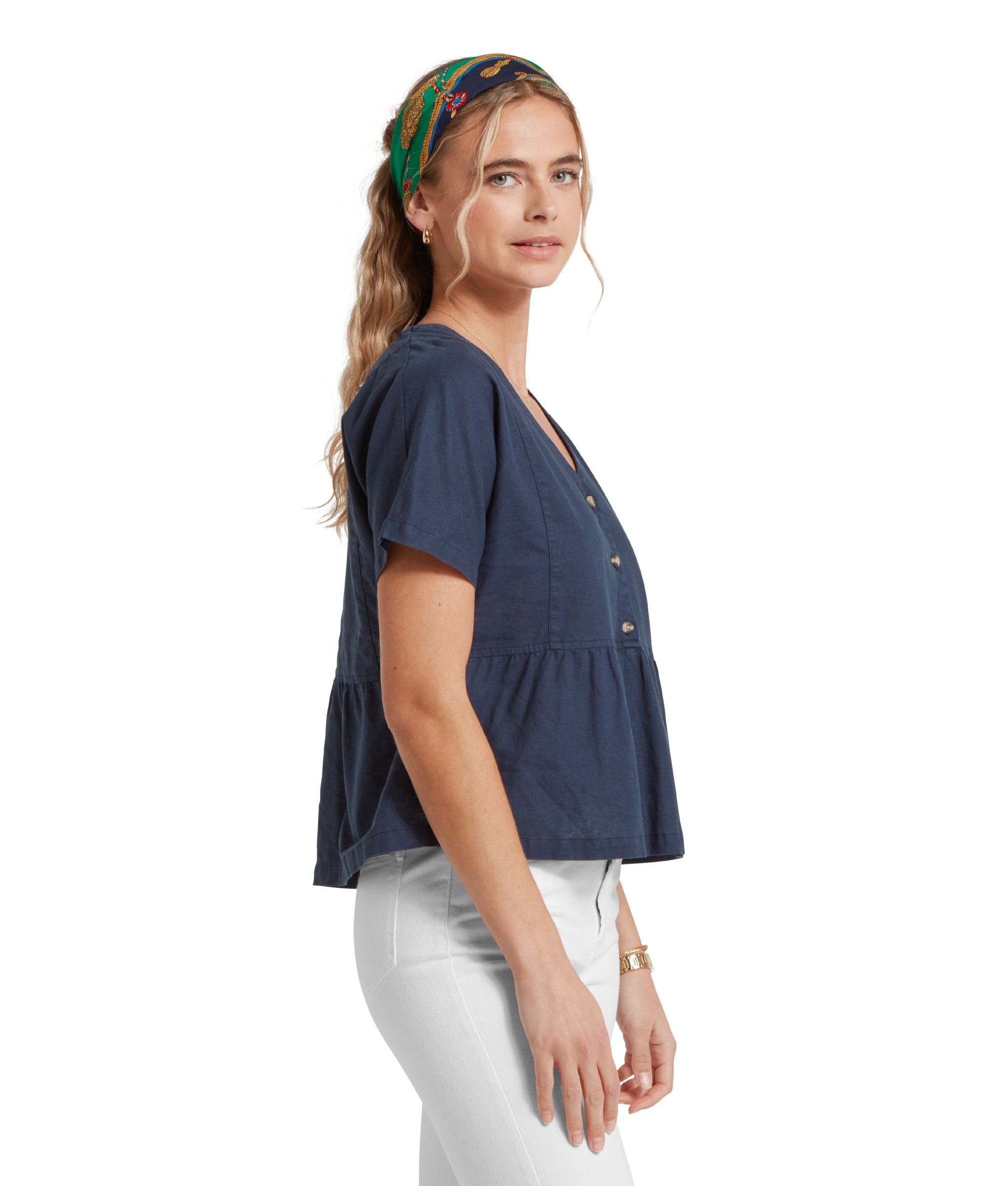 Side profile image of a female model wearing a v-neck navy linen Schöffel top, paired with white jeans. She is standing in a relaxed pose with her hands by her side.