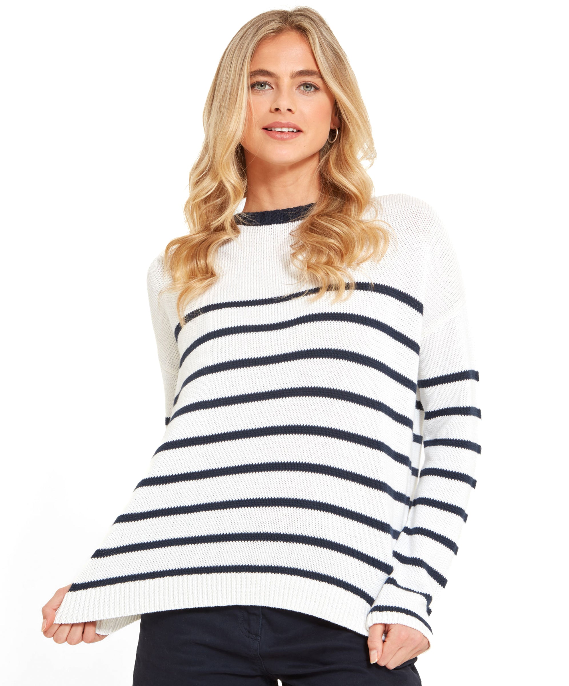 A blonde woman is wearing a white jumper with navy stripes, posing while gently pulling the hem of the jumper.