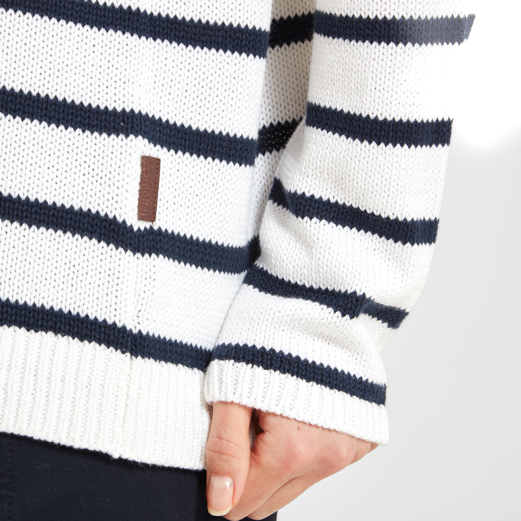 A close-up of the hem of the white jumper with navy stripes, highlighting a small brown leather tag with branding.