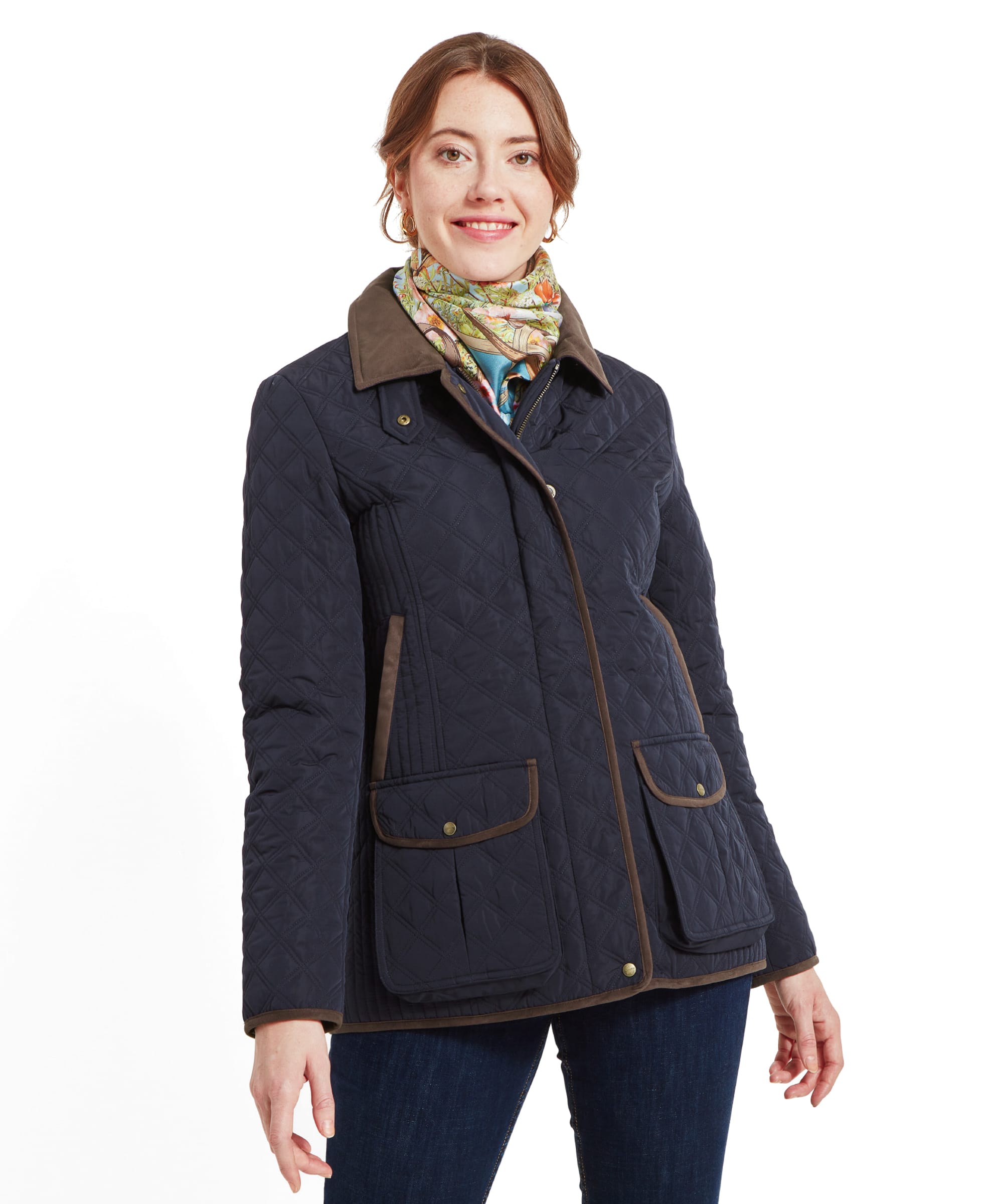 A woman wearing a Schöffel Lilymere Quilted Jacket for Women in Blue with a brown collar and two front pockets. She is also wearing a patterned scarf and dark jeans. The jacket is buttoned up, and she is smiling slightly.