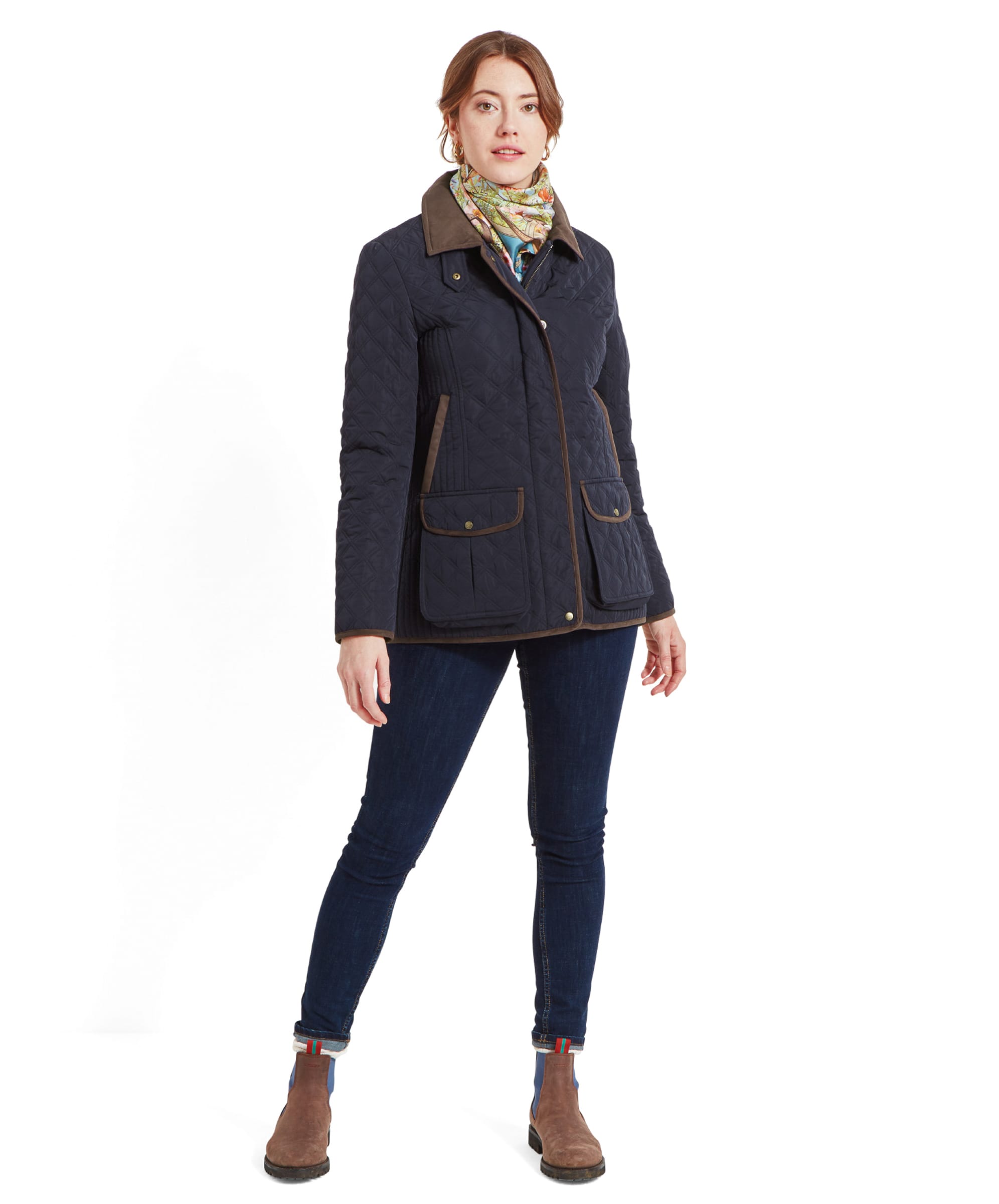 Full body shot of the woman in the Schöffel Lilymere Quilted Jacket for Women in Blue, brown collar, and scarf. She is standing with her hands by her sides, wearing dark jeans and ankle boots.