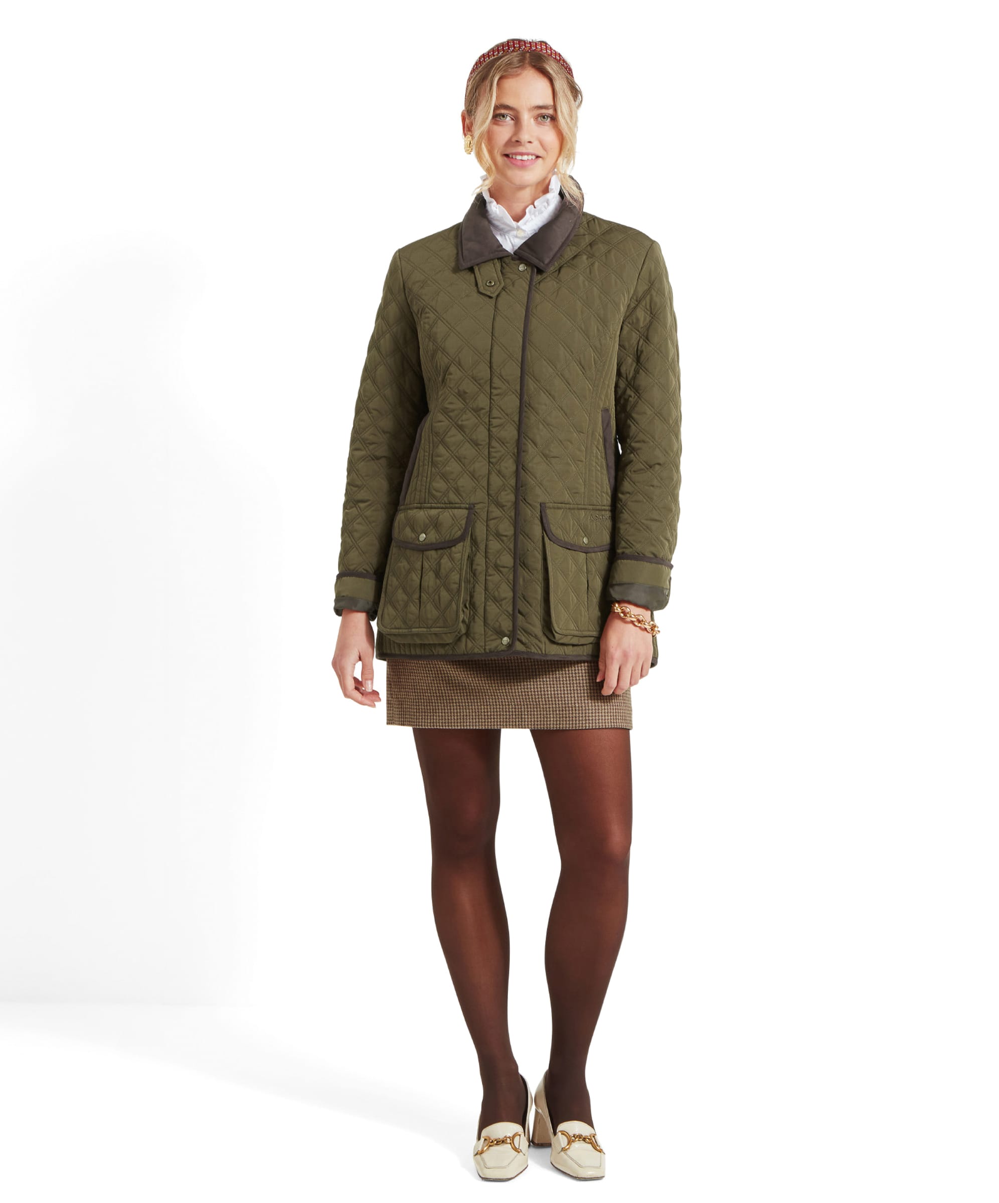 The woman is standing in the Schöffel Lilymere Quilted Jacket for Women in Green, which has brown trim and a dark collar. She pairs the jacket with a houndstooth mini skirt, black tights, and cream-colored loafers. Her pose is full-body, showing the outfit completely.