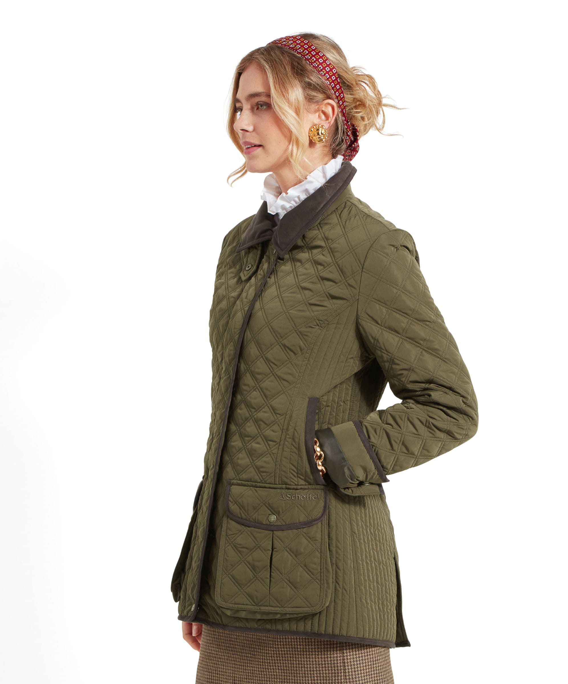 The woman is seen standing in a three-quarter view, showcasing the Schöffel Lilymere Quilted Jacket for Women in Green with a dark collar and brown trim. The jacket has two front flap pockets, and the woman’s hands are positioned near the front of the jacket, as if holding it closed.