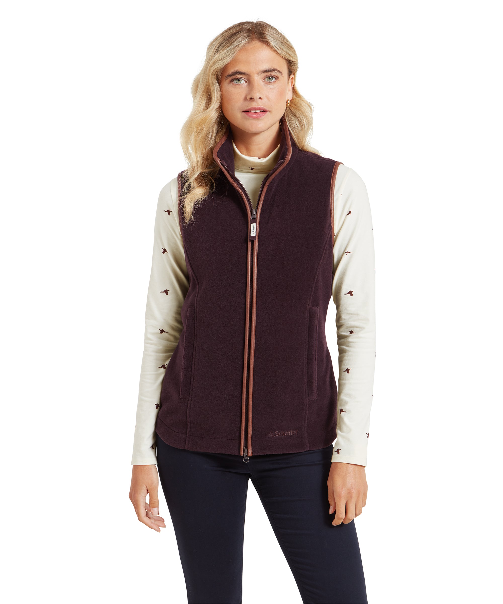 A woman is wearing a Schöffel Lyndon Fleece Gilet for Women in Brown over a cream long-sleeved top with bird patterns. The gilet features a high collar, a full-length zip, and side pockets. She stands with a neutral expression, looking directly at the camera.