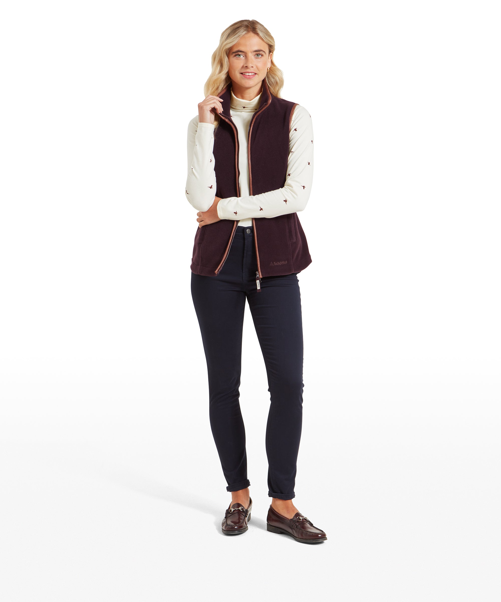 The same woman is shown in a full-body view wearing the Schöffel Lyndon Fleece Gilet for Women in Brown, paired with dark jeans and brown loafers. She is standing straight with one hand lightly holding the collar of the gilet, smiling.