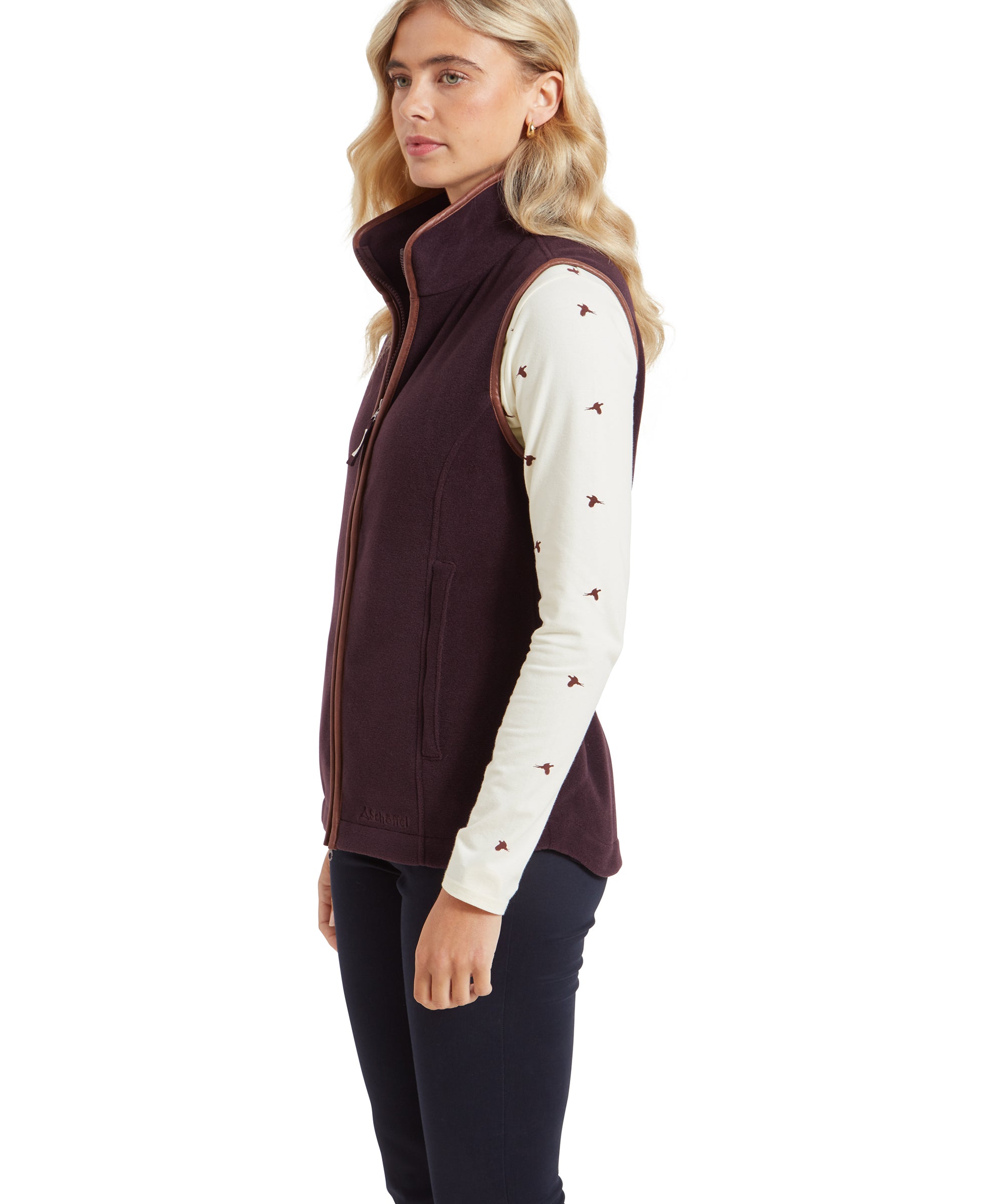 The side profile of the woman highlights the slim fit of the Schöffel Lyndon Fleece Gilet for Women in Brown. The high collar and zippered side pockets are visible, with the bird pattern on the cream top showing clearly under the gilet.