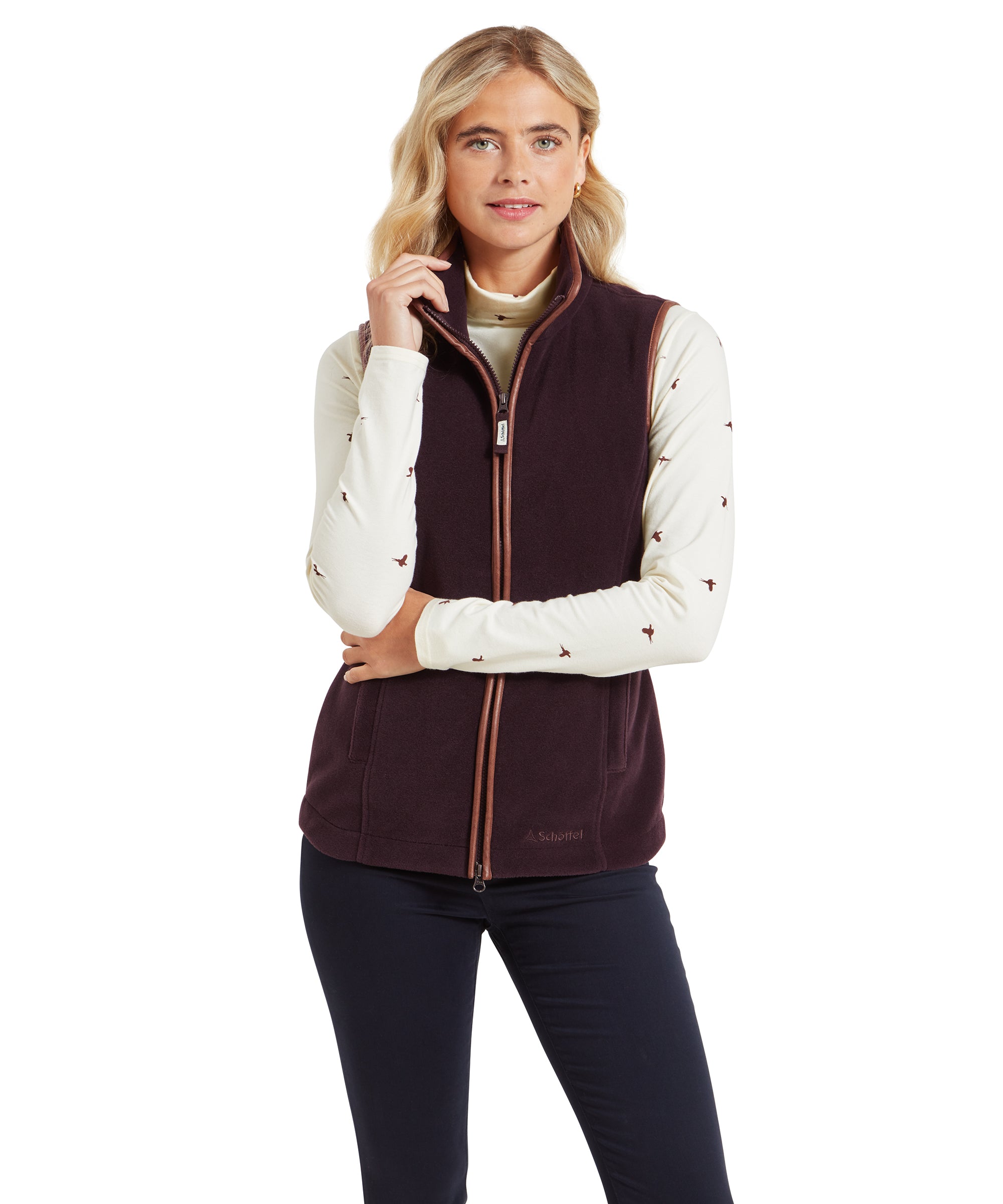 A woman is wearing a Schöffel Lyndon Fleece Gilet for Women in Brown over a cream long-sleeved top with a bird pattern. The gilet has a high collar and a full-length zip with contrasting trim. The woman is standing with one hand lightly holding the collar of the gilet, and she is smiling softly while looking directly at the camera. She is also wearing dark trousers, completing the look with a casual yet polished style.