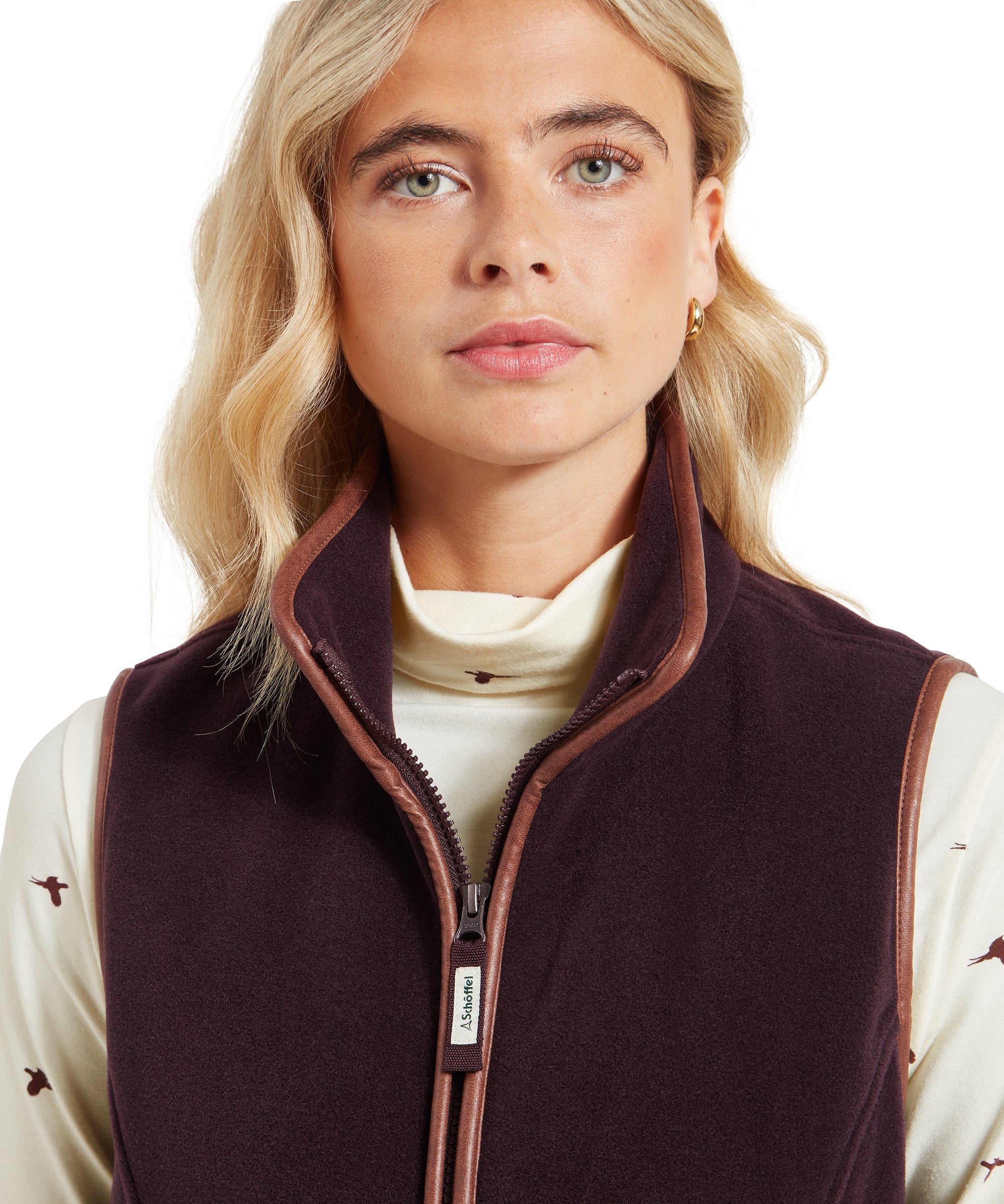 A detailed close-up of the woman’s face and the top of the Schöffel Lyndon Fleece Gilet for Women in Brown, focusing on the high collar, zip pull, and the contrast trim along the edges. The cream top with the bird pattern is partially visible above the collar.