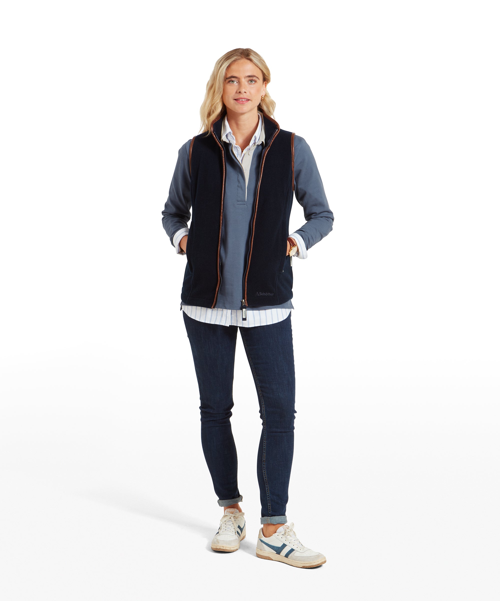 The same woman is shown in a full-body shot, standing with her hands in her pockets. She wears the Schöffel Lyndon Fleece Gilet for Women in Navy, blue jeans, and brown boots. The gilet's brown trim complements the casual yet polished look.