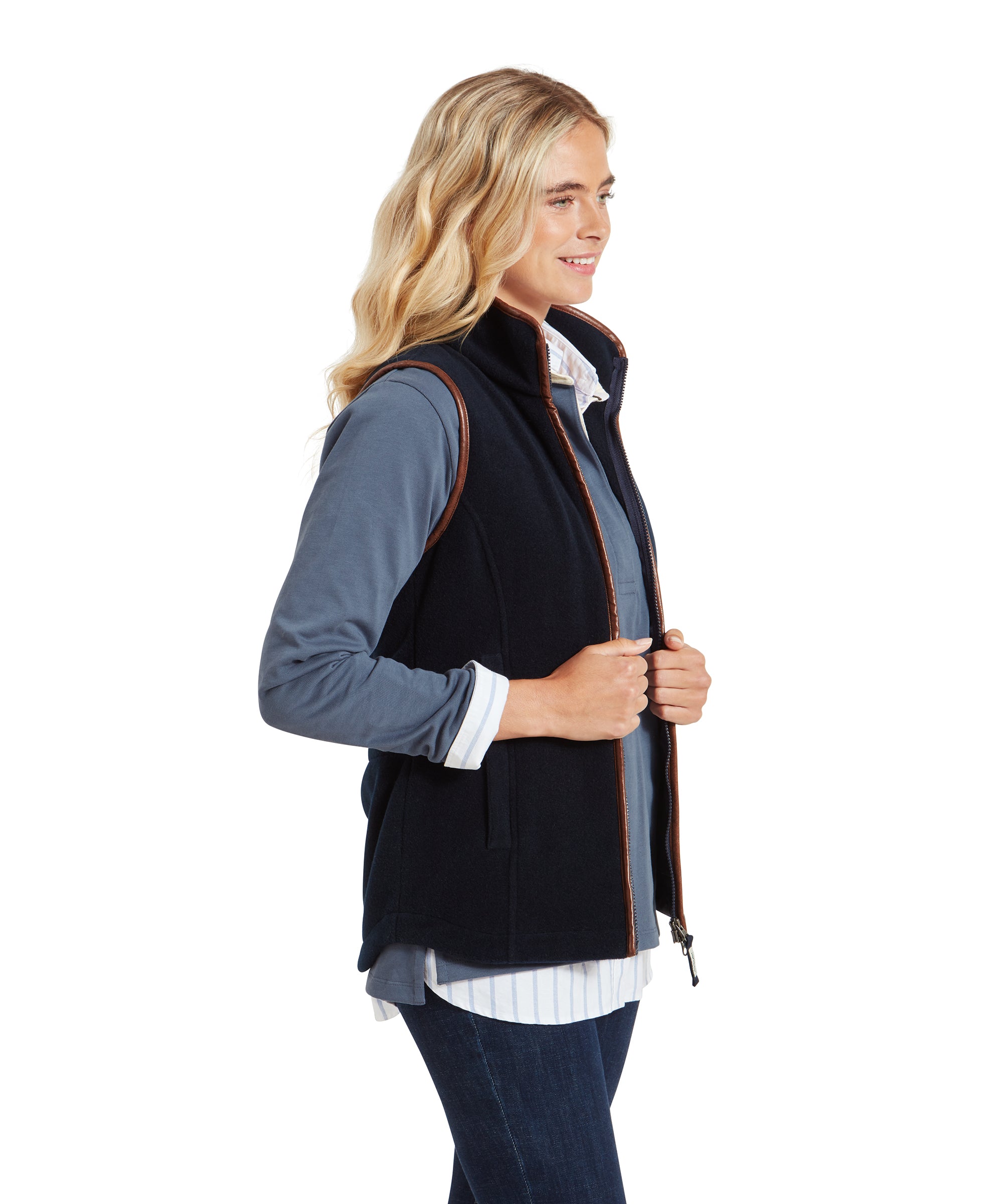 The same woman is shown in a full-body shot, standing with her hands in her pockets. She wears the Schöffel Lyndon Fleece Gilet for Women in Navy, blue jeans, and brown boots. The gilet's brown trim complements the casual yet polished look.