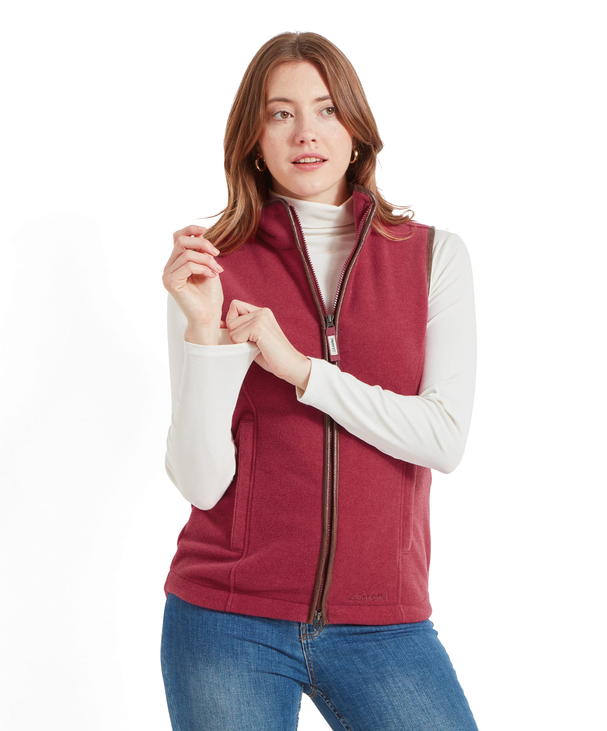 A woman wearing a Schöffel Lyndon Fleece Gilet for Women in Red. The gilet is sleeveless with a full front zip, and she is posing with her hands near the zipper. She is wearing a white turtleneck underneath and blue jeans.