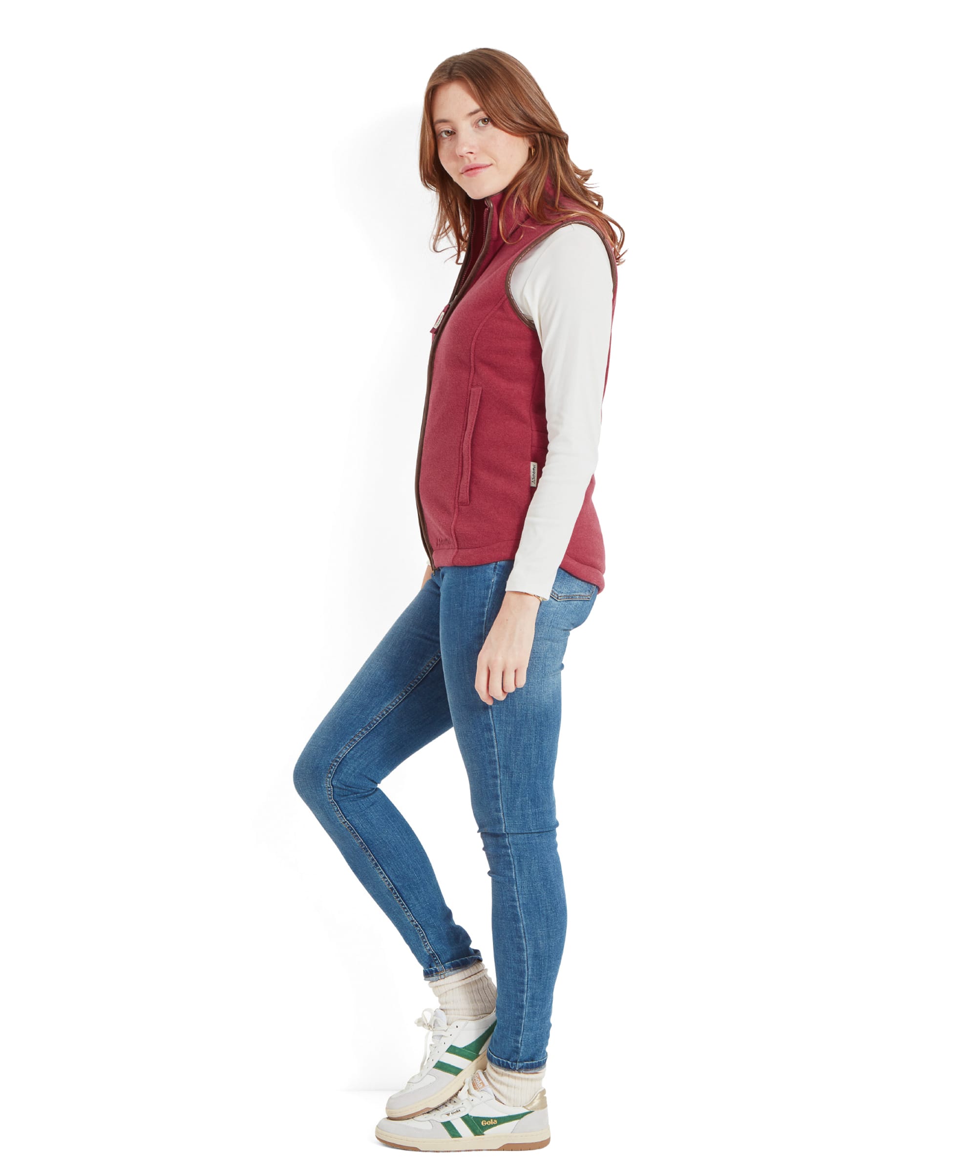 The same woman is standing in a casual pose, wearing a Schöffel Lyndon Fleece Gilet for Women in Red over a white turtleneck, paired with blue jeans and white trainers with green accents. She has one hand in her hair, looking down with a smile.