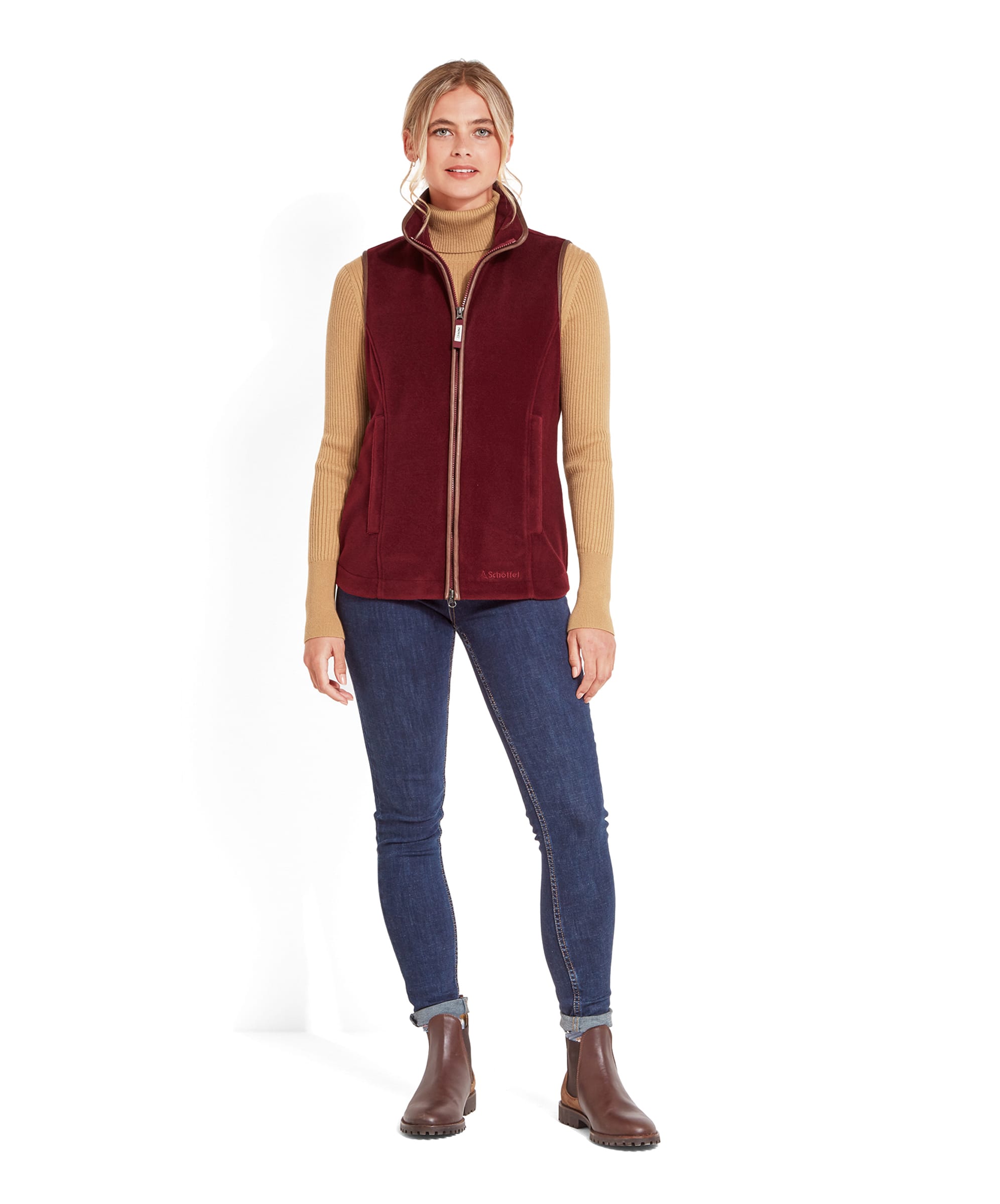 The same woman wearing the Schöffel Lyndon Fleece Gilet for Women in Deep Red, standing with her legs slightly apart. She is also wearing dark blue jeans and brown ankle boots, with her hands rested to her sides.