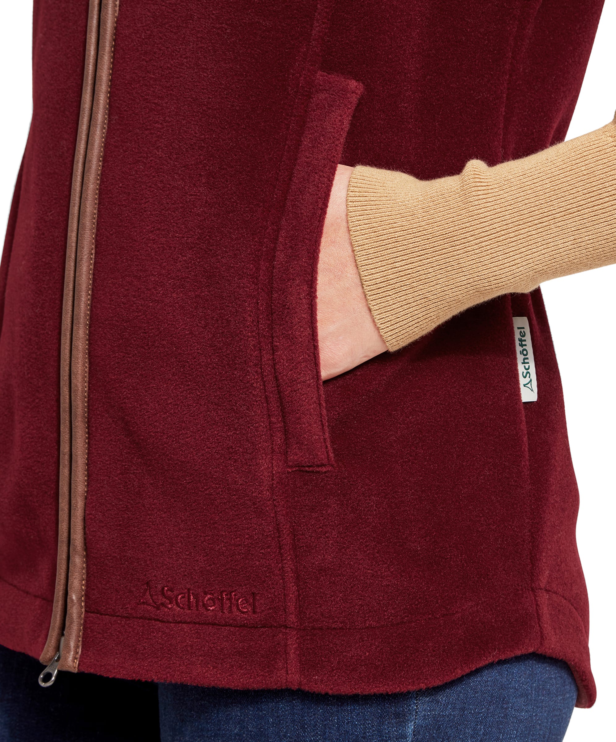 A close-up of the Schöffel Lyndon Fleece Gilet for Women in Deep Red, focusing on the pocket and the Schöffel logo embroidered near the hem. The woman's hand is tucked into the pocket.