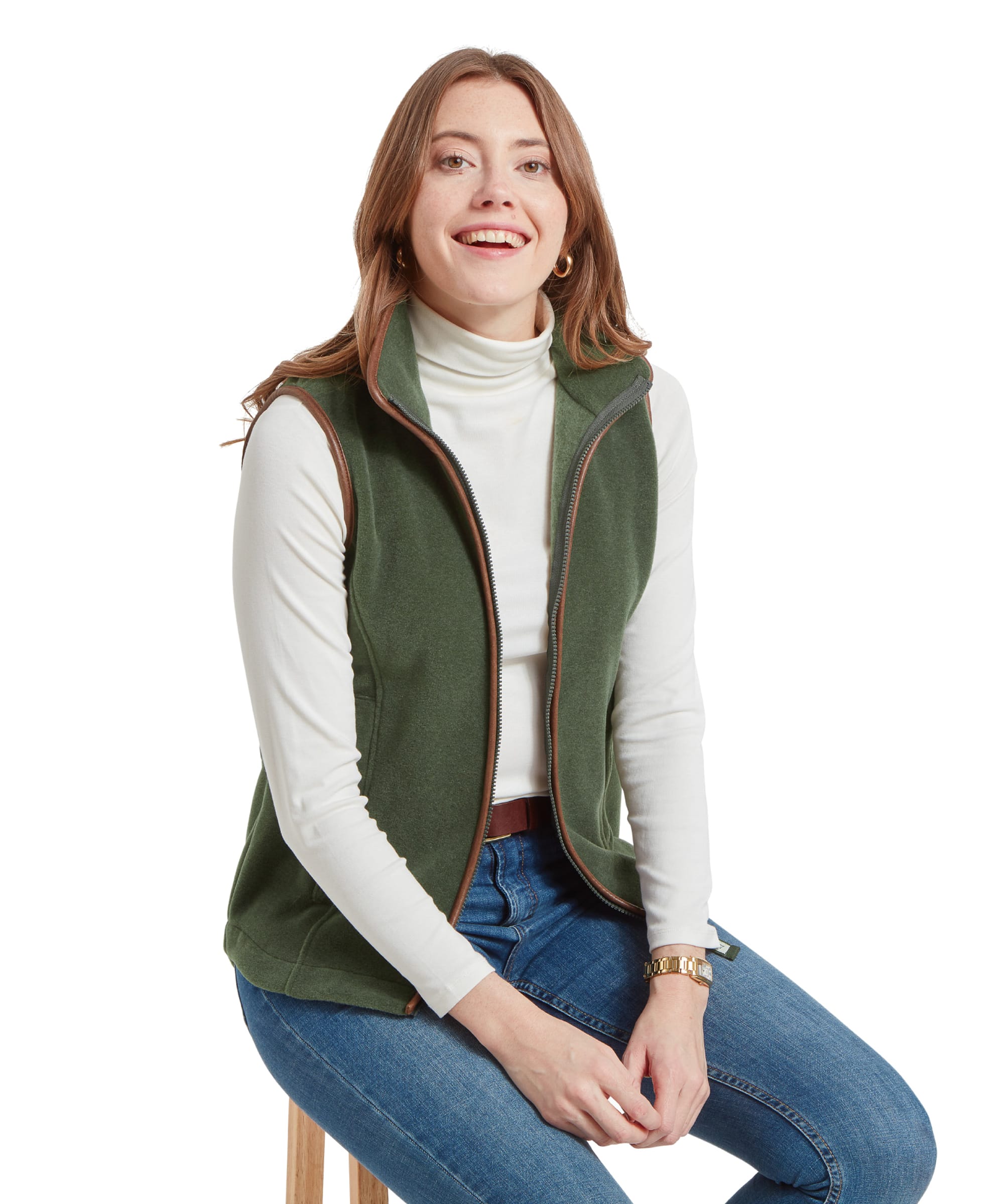 Woman sitting on a stool, smiling, wearing a Schöffel Lyndon Fleece Gilet for Women in Green over a white turtleneck and blue jeans.