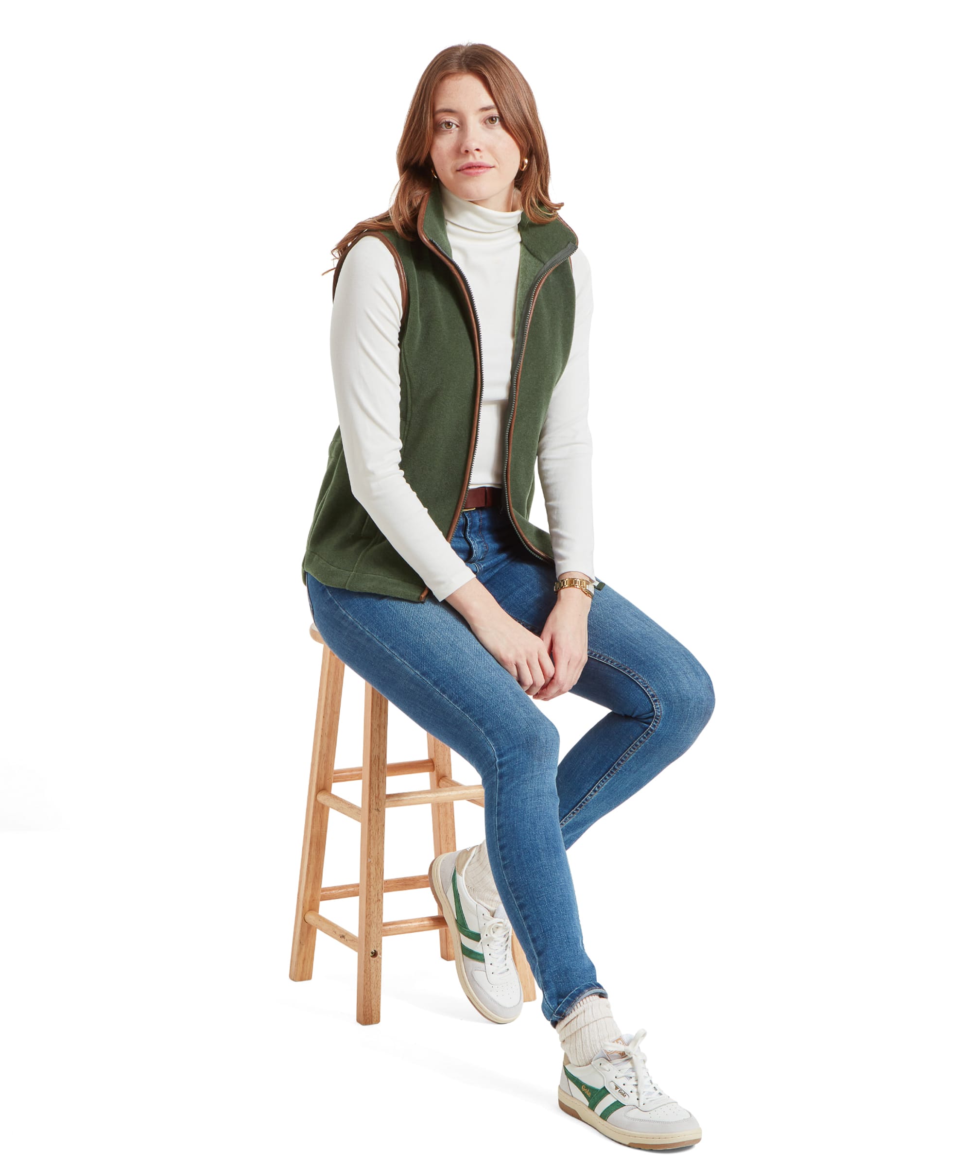 Full-body view of a woman sitting on a stool, wearing a Schöffel Lyndon Fleece Gilet for Women in Green over a white turtleneck, blue jeans, and white sneakers with green stripes.