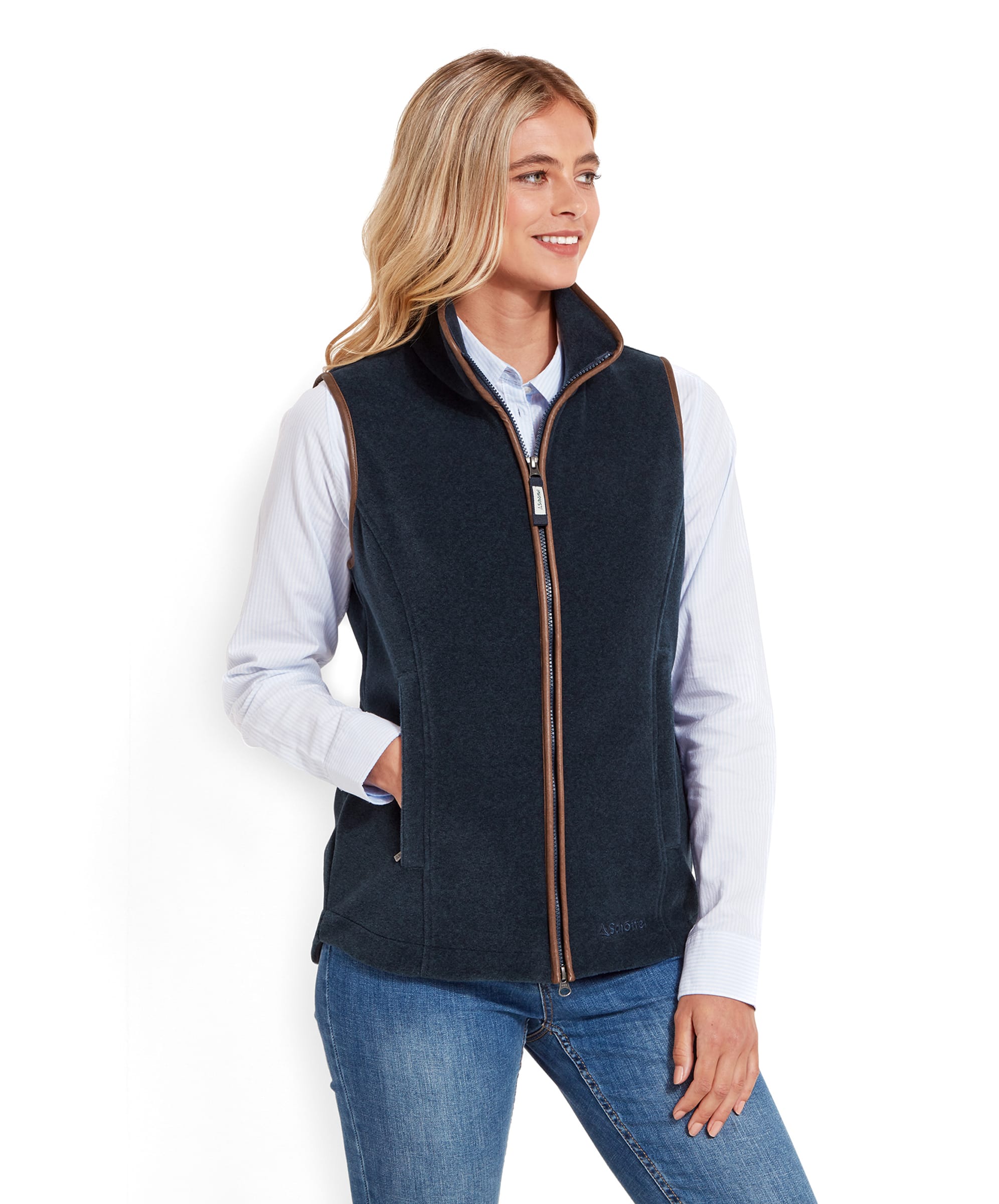 A woman is wearing a Schöffel Lyndon Fleece Gilet for Women in Navy Blue over a light blue button-up shirt. The gilet has brown trim along the edges and a zip closure. She stands with one hand in her pocket, smiling slightly, with long blonde hair flowing down her shoulders.