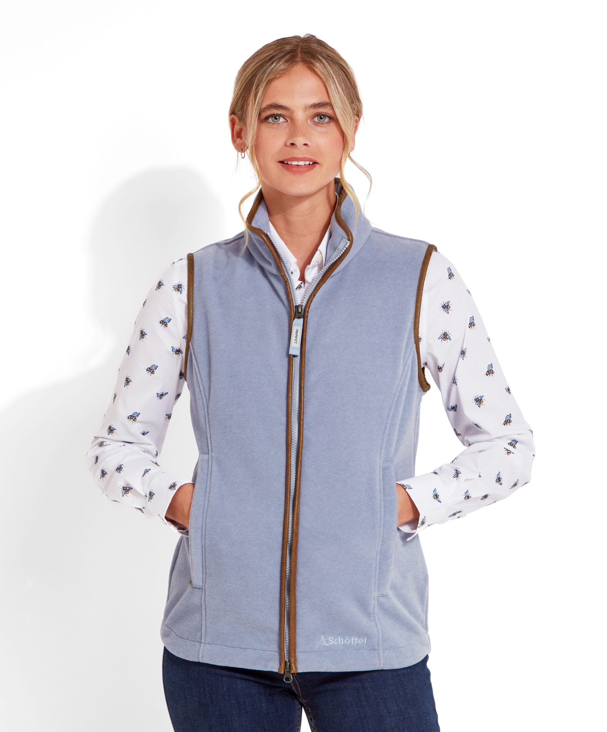 Woman wearing a Schöffel Lyndon Fleece Gilet for Women in Pale Blue with a white shirt that has bee patterns. The gilet has a brown trim and a high collar. The woman is standing with her hands in the pockets, smiling slightly at the camera.