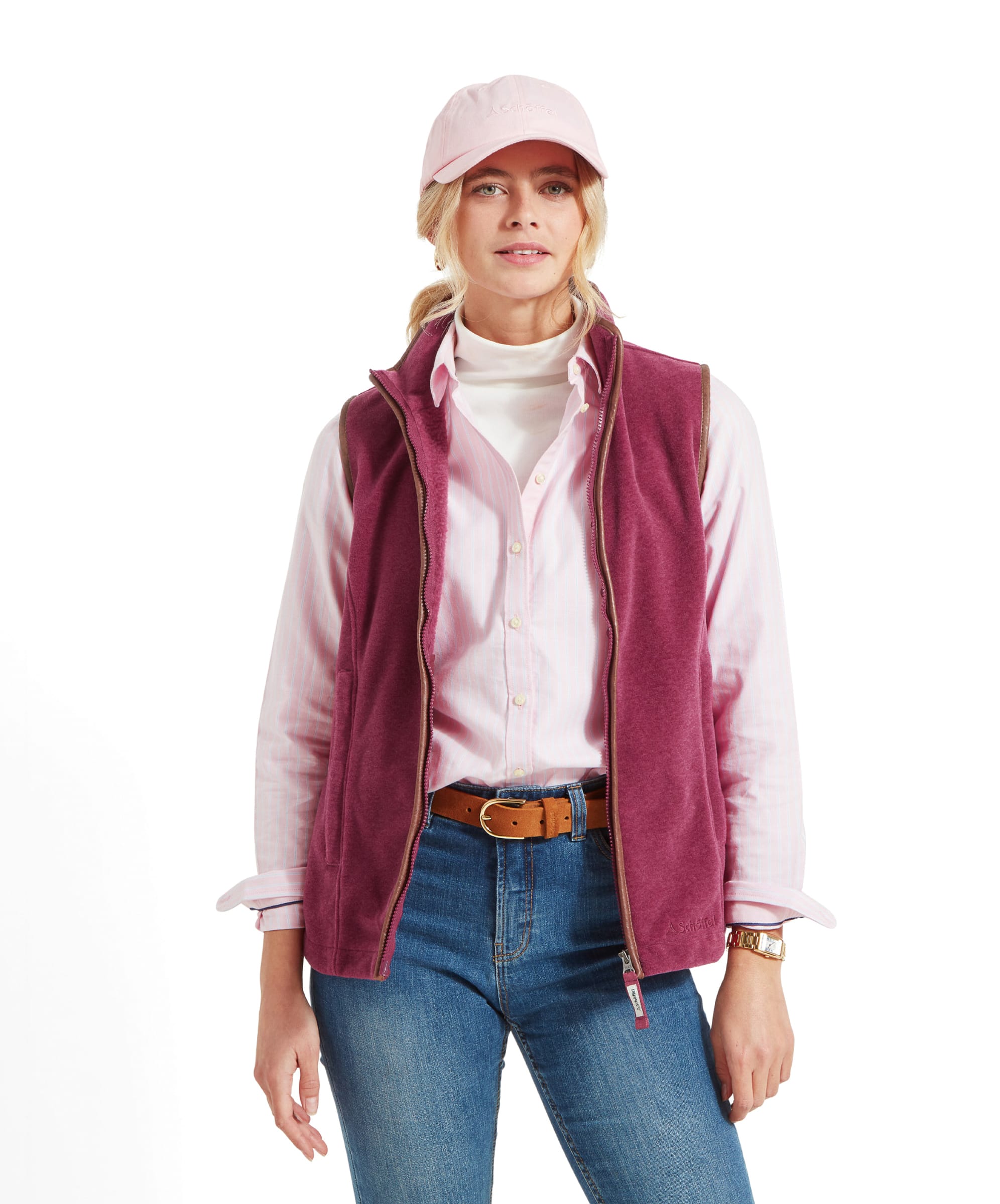 A woman wearing a pale pink shirt and Schöffel Lyndon Fleece Gilet for Women in Red, with a matching pale pink cap. She is standing with her hands in the pockets of the gilet, which has a brown leather trim along the zipper and armholes. She is wearing light blue jeans and a brown leather belt.