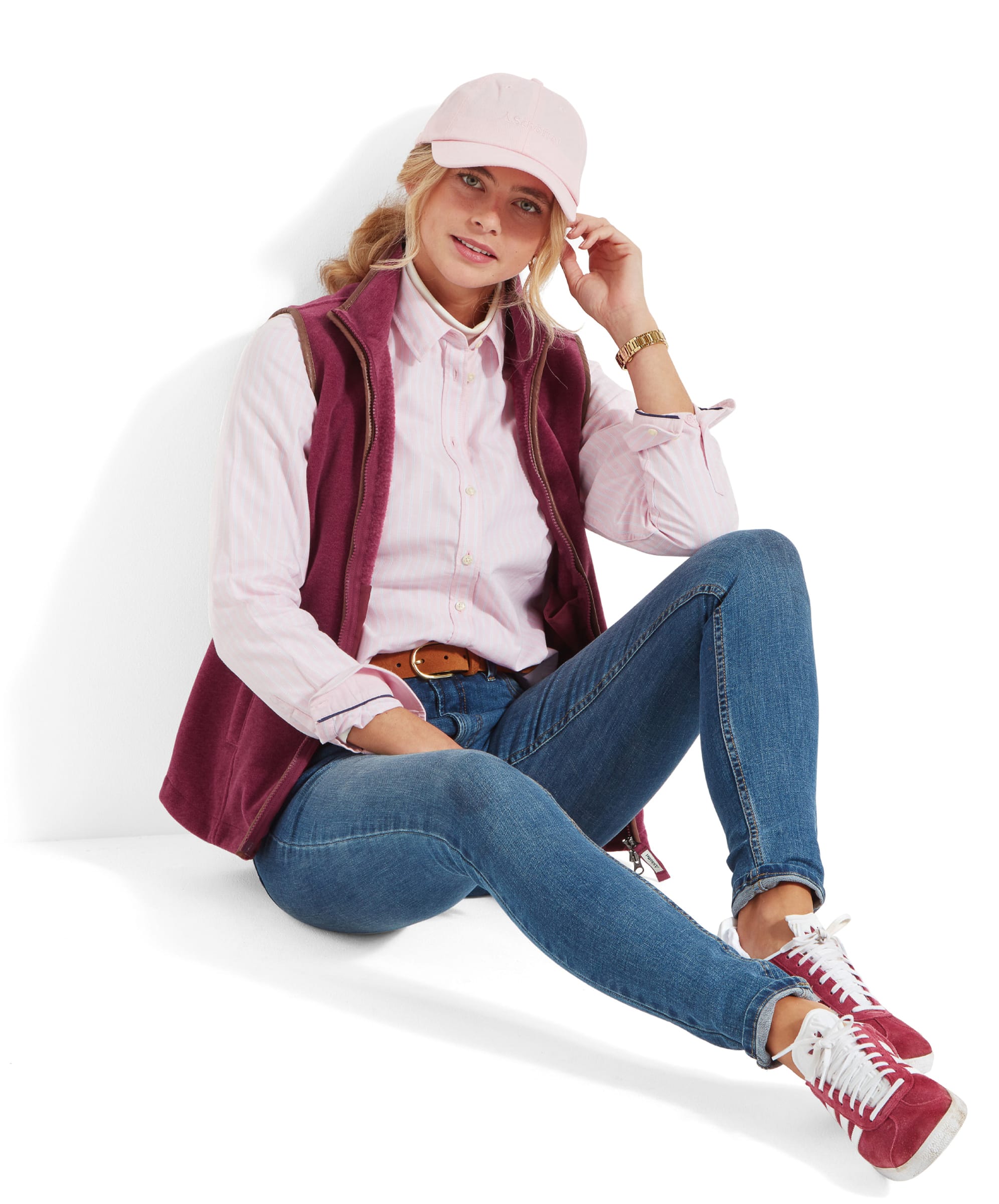 The same woman sitting on the floor against a white background, wearing a pale pink shirt, Schöffel Lyndon Fleece Gilet for Women in Red, and pale pink cap. She has her right leg bent and her left leg stretched out, resting her elbow on her knee while adjusting her cap. She pairs her outfit with light blue jeans, a brown leather belt, and burgundy and white trainers.