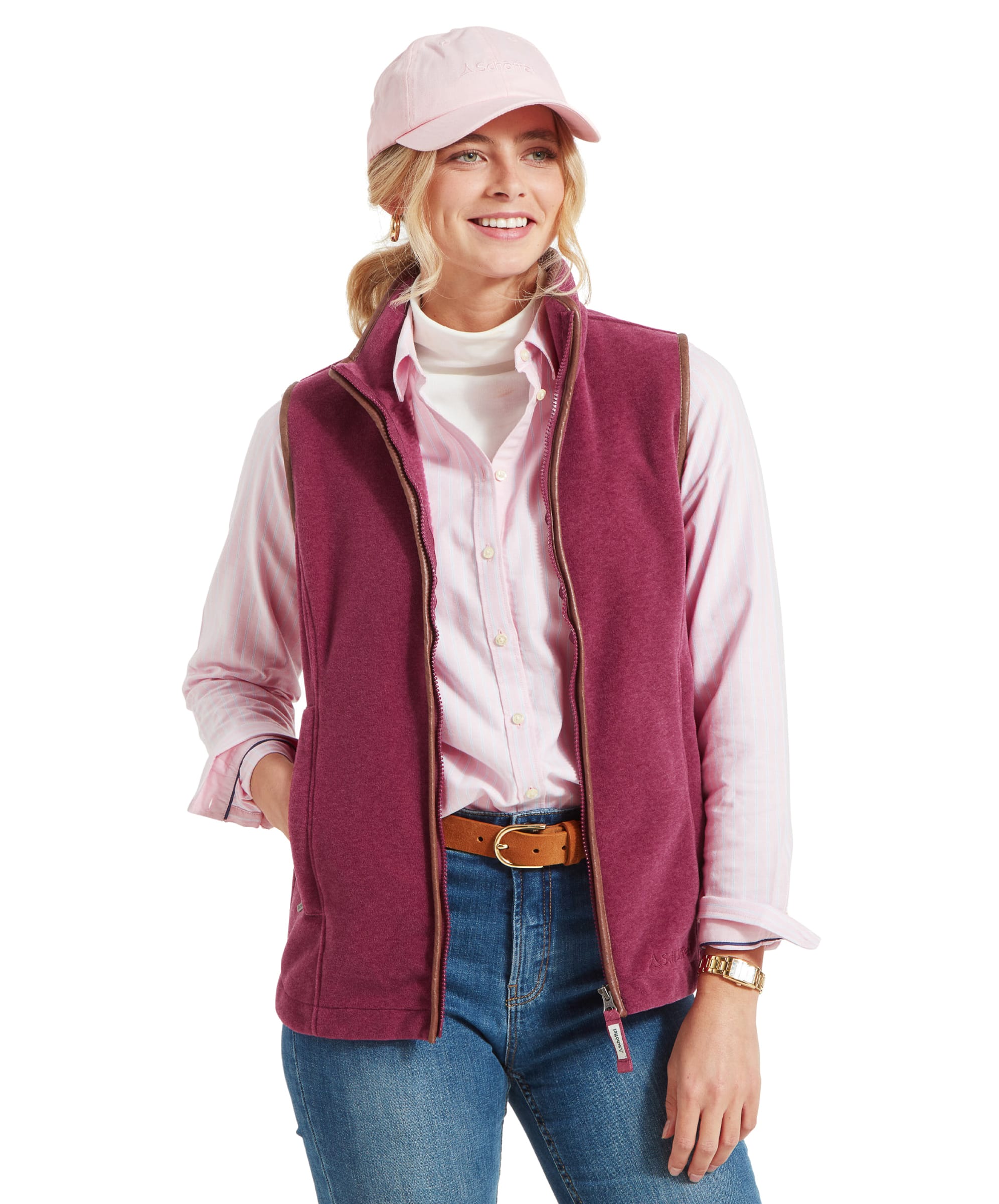 The same woman is standing and smiling, wearing the Schöffel Lyndon Fleece Gilet for Women in Red, pale pink shirt, and pale pink cap. She is resting her left hand in her gilet pocket.