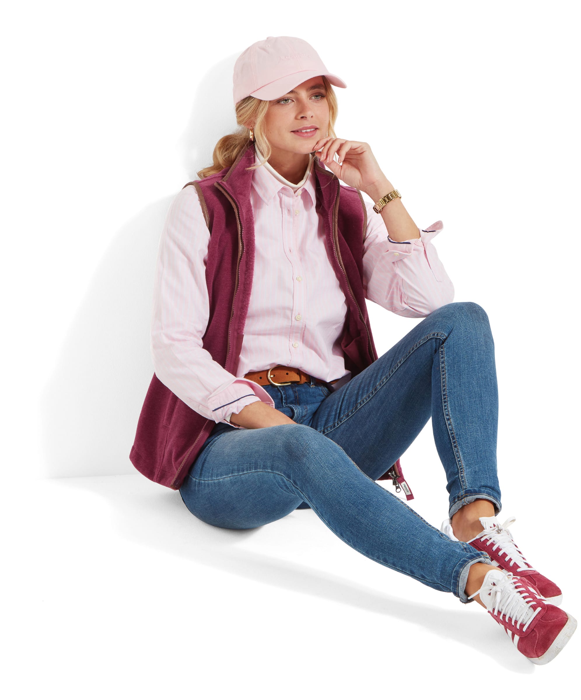 A woman wearing a Schöffel Lyndon Fleece Gilet for Women in Red, a pale pink shirt, and a pale pink cap sits casually on the floor with her back resting against a white wall. She has her left leg bent and her right leg extended, resting her left elbow on her knee while holding her chin with her left hand. She is also wearing light blue jeans, a brown leather belt, and burgundy trainers with white laces. She is smiling softly and looking slightly to her right.