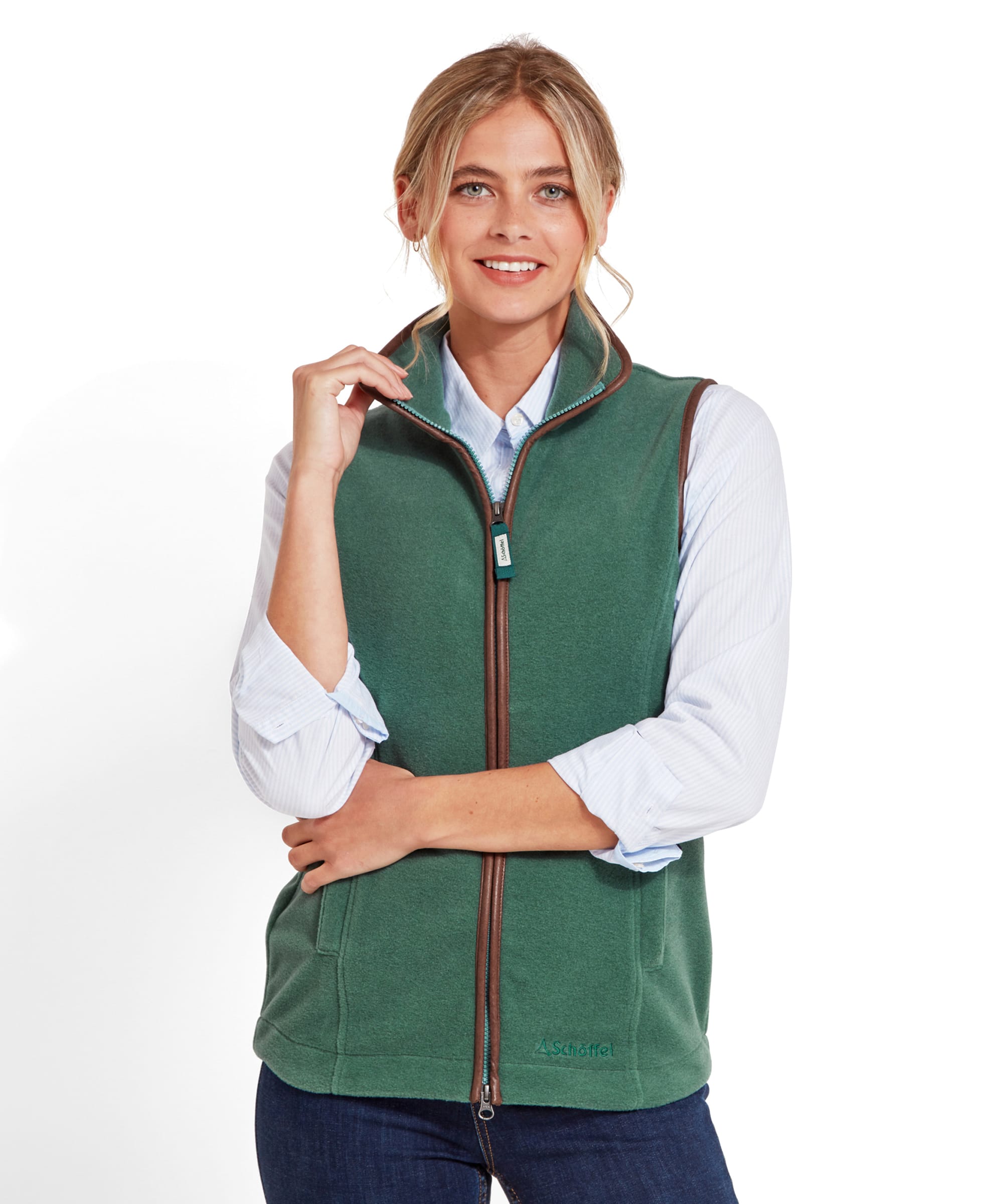 A woman wearing a Schöffel Lyndon Fleece Gilet for Women in Teal Blue with brown leather trim over a light blue collared shirt. The gilet features a zippered front and side pockets, and the woman is smiling, with one hand adjusting the collar.