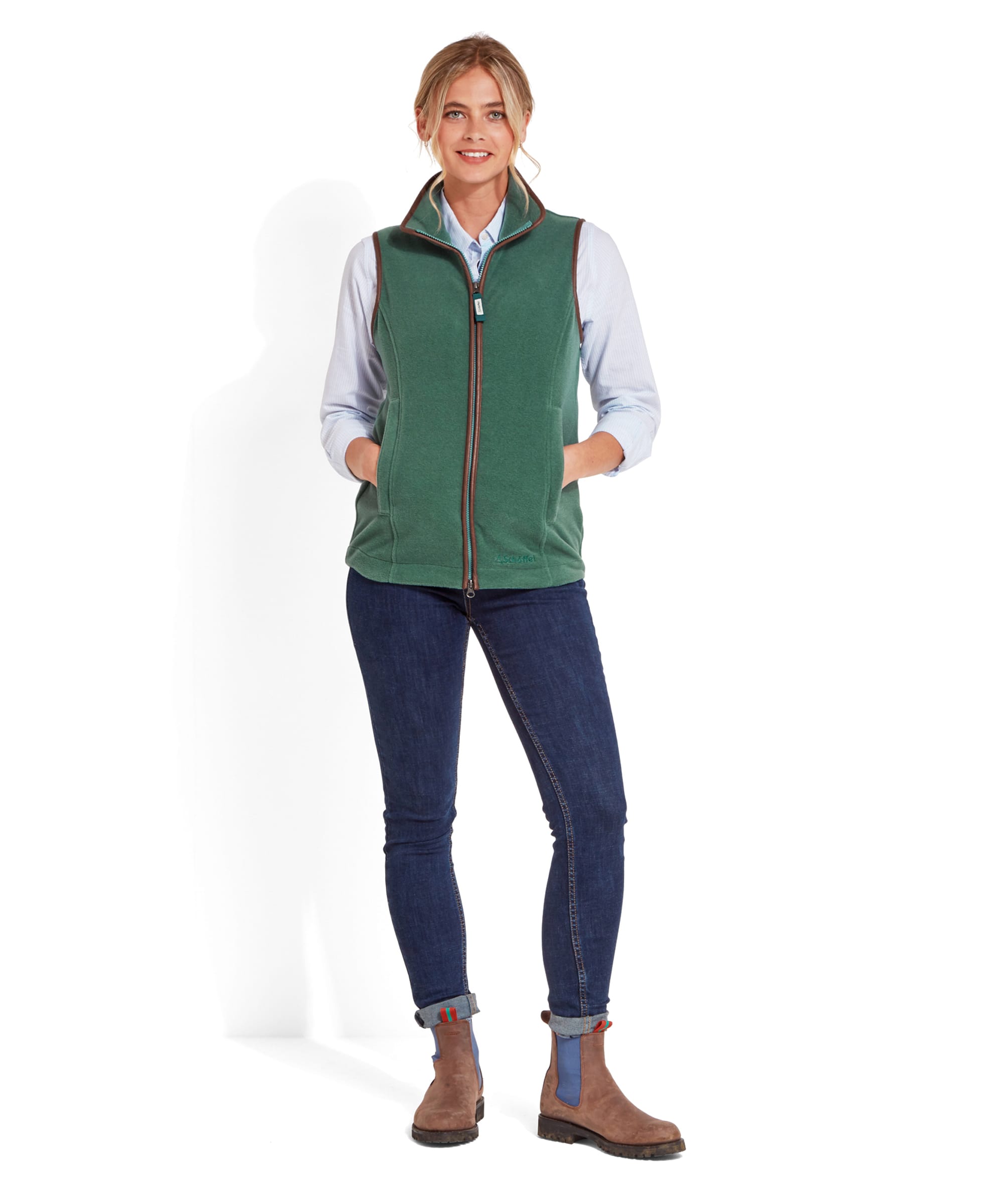 Full-body shot of the same woman in a Schöffel Lyndon Fleece Gilet for Women in Teal Blue with brown trim, paired with a light blue shirt, dark blue jeans, and brown ankle boots. She stands with her hands in her pockets, smiling.