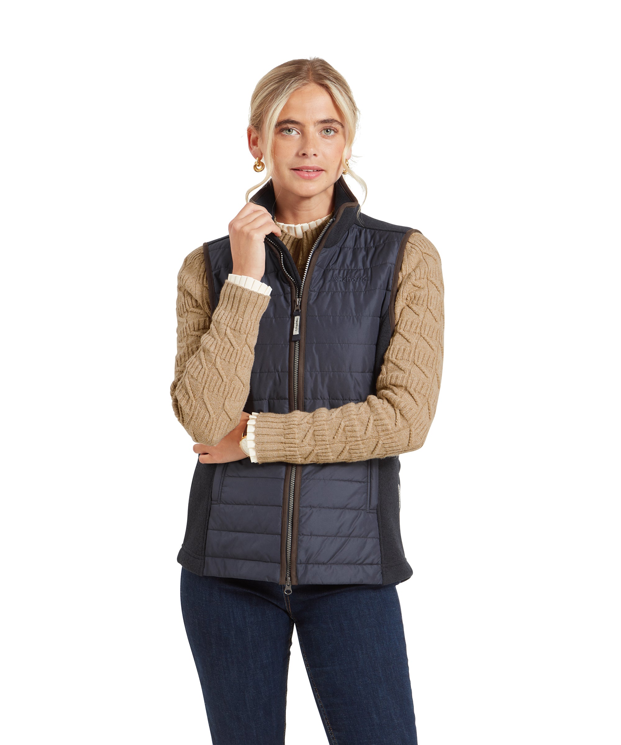 A woman wearing a Schöffel Lyndon Hybrid Gilet for Women in Blue with brown trim over a tan knitted sweater. She is standing with one hand on the collar of the gilet, smiling slightly. She has blonde hair pulled back and is wearing hoop earrings.